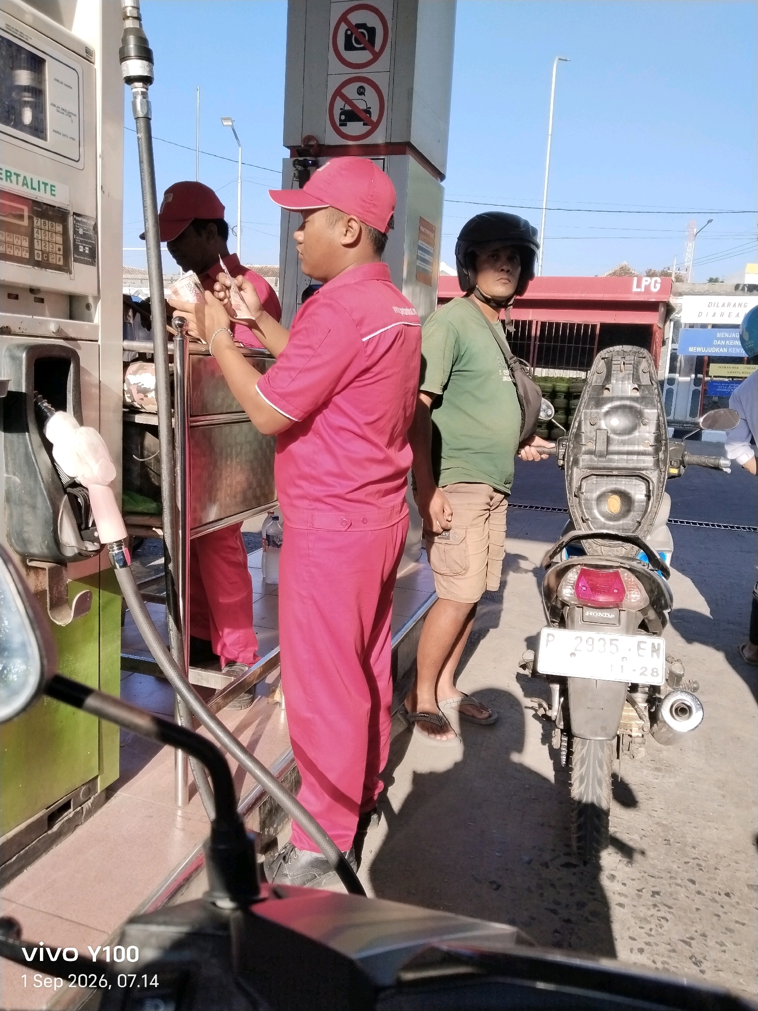 The image captures a scene at a gas station in Situbondo, Indonesia, on September 1, 2026, at 07:14. The focus is on a gas pump and several individuals. In the foreground, a motorcycle is partially visible, with its seat and rear fender featuring a license plate that reads "P 2935 EN" and "11-28".
To the right of the gas pump, a person wearing a green t-shirt and beige cargo shorts stands with a black helmet under their arm, looking towards the camera. They have a dark bag slung over their shoulder. The gas pump itself is silver and green, with a nozzle hanging out.
To the left of the gas pump, two individuals in red uniforms and matching red caps are interacting. One person, facing away from the camera and slightly blurred, appears to be holding and counting Indonesian Rupiah banknotes. The other person, facing more towards the camera, is also in a red uniform and appears to be observing or assisting. They are standing near the gas pump console.
In the background, there is a red building with the letters "LPG" visible, along with some signage. The sky is clear and blue. The overall environment suggests an outdoor, public space. The image includes a watermark in the bottom left corner: "vivo Y100 1 Sep 2026, 07.14".