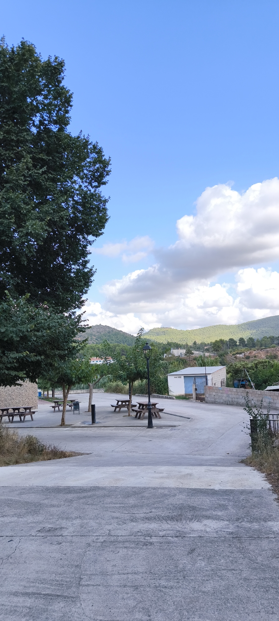 This image shows a public park area with picnic tables and benches in Fanzara, Spain. A large tree is prominent on the left side of the frame, with its foliage extending towards the bright blue sky with scattered white clouds. In the background, rolling green hills with scattered buildings can be seen. The park area itself is paved with concrete and features several wooden picnic tables with attached benches. A black lamppost stands centrally in the park. To the right, there is a small white building with a blue-paneled door and a brick wall. The scene appears to be during daylight, under a clear sky with some cloud cover.