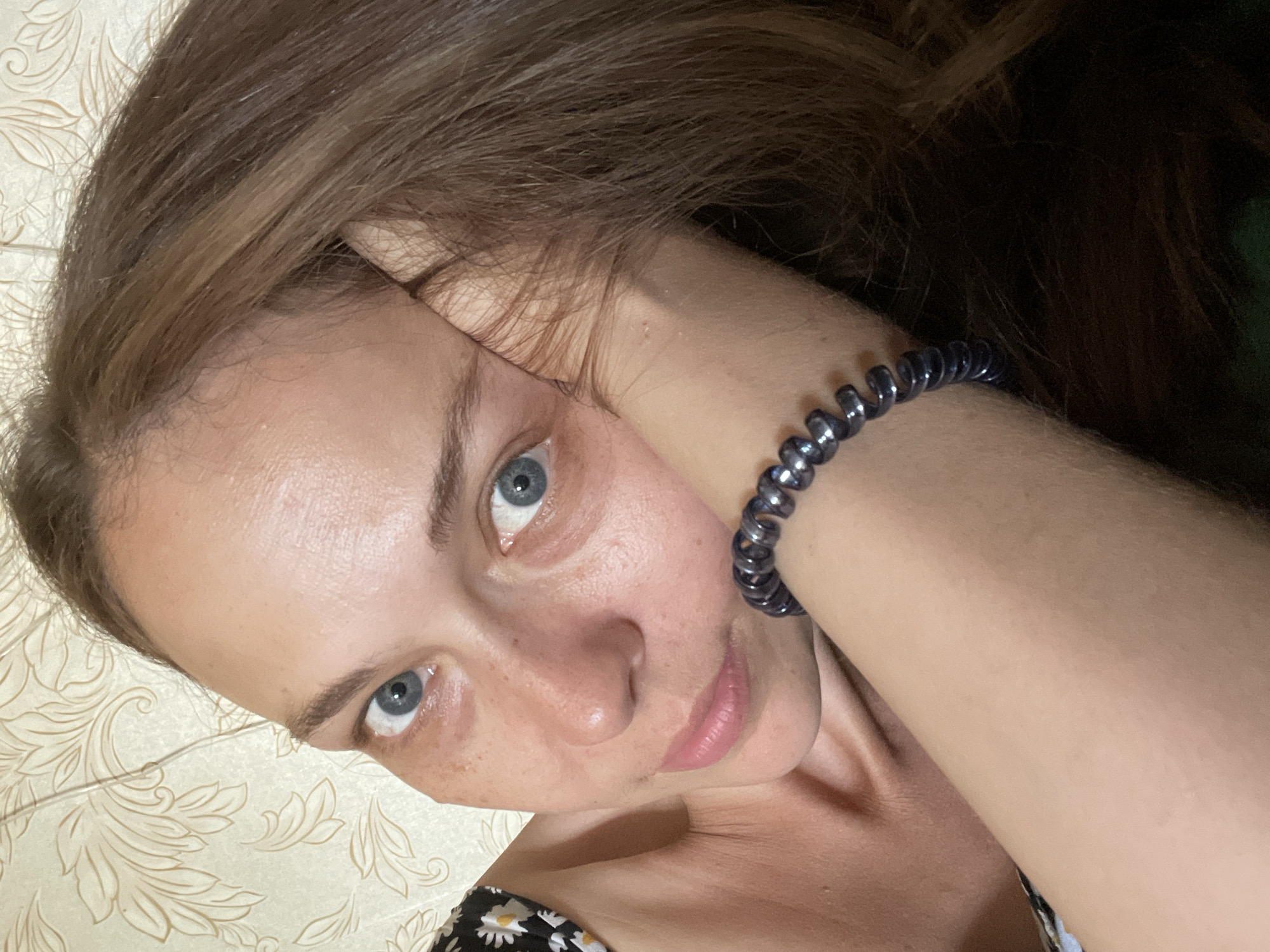 A close-up selfie shows a young woman with brown hair and blue eyes resting her head on her hand. She is wearing a dark bracelet with metallic beads on her wrist. Her skin has a smattering of freckles. The background appears to be a wall with a floral pattern. The lighting is soft and natural. The image is cropped to focus on the woman's face and upper body.