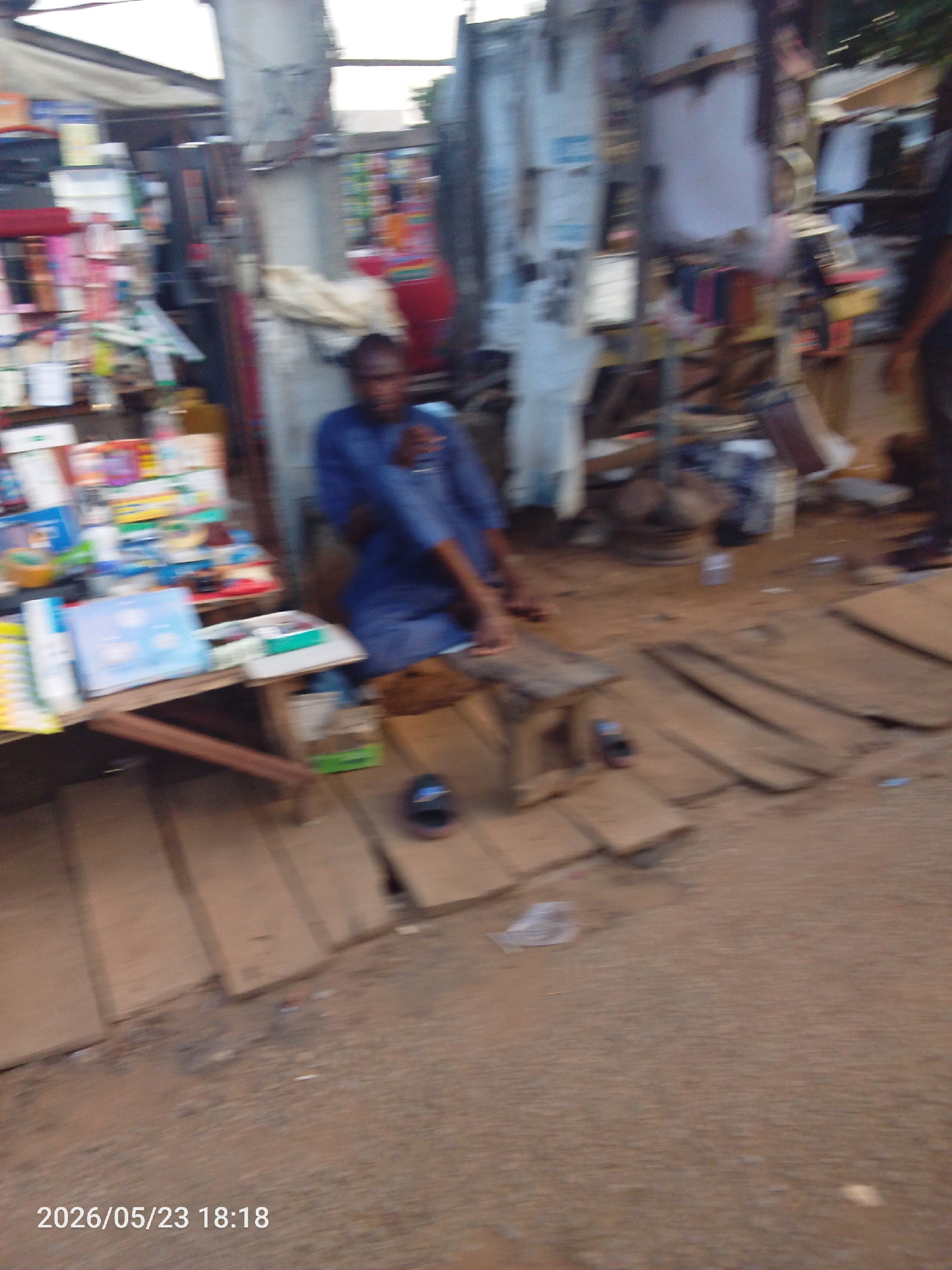 An image of an outdoor street scene, possibly a market in Lokoja, Nigeria. An individual is seated on a low wooden stool, wearing what appears to be a blue garment. The person is next to a makeshift table with books and other items for sale. The ground is covered with wooden planks and dirt. Additional merchandise is visible in the background, indicating a trading environment.
