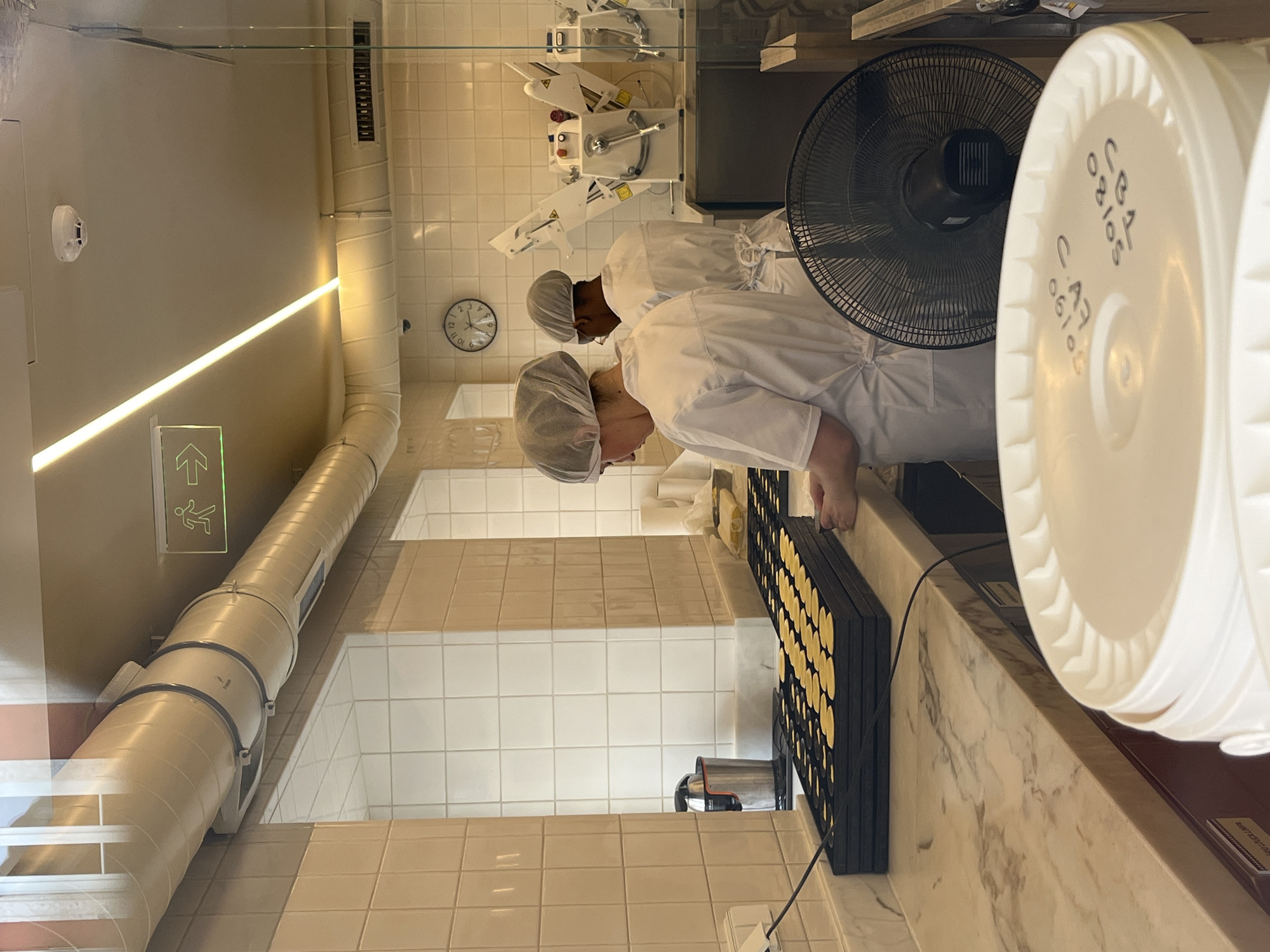 In a commercial bakery setting in Lisbon, Portugal, two individuals wearing white chef coats and hairnets are actively engaged in preparing what appears to be small, round pastries. The scene includes a white-tiled wall behind the workers, a complex of machinery associated with baking or food processing in the background, and a large black electric fan positioned to the side. A rack filled with circular, yellowish pastries is visible on a marble countertop, where the workers are focused on their task. The environment is illuminated by an overhead fluorescent light, and near the ceiling is a sign with a green background, possibly indicating an exit or directional information. A large, white container with handwritten text is partially obscuring part of the scene.