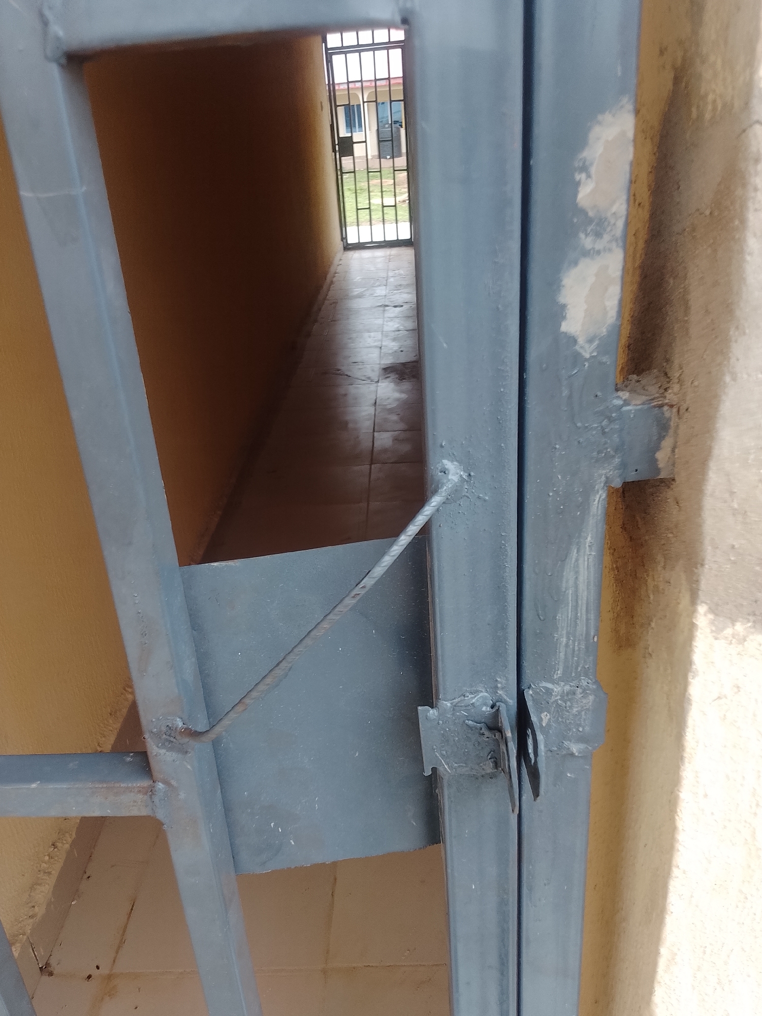 The image shows a close-up of a grey metal gate and frame, with a narrow hallway visible in the background. The hallway is lined with a light brown wall and a tiled floor. At the end of the hallway, a black metal gate with vertical bars is visible, and beyond that, a building with windows and a grassy area can be seen. The metal gate in the foreground appears to be attached to a textured, light-colored wall on the right side of the image. There is a thin rope or cable attached to the gate and extending into the hallway. The scene is outdoors.
