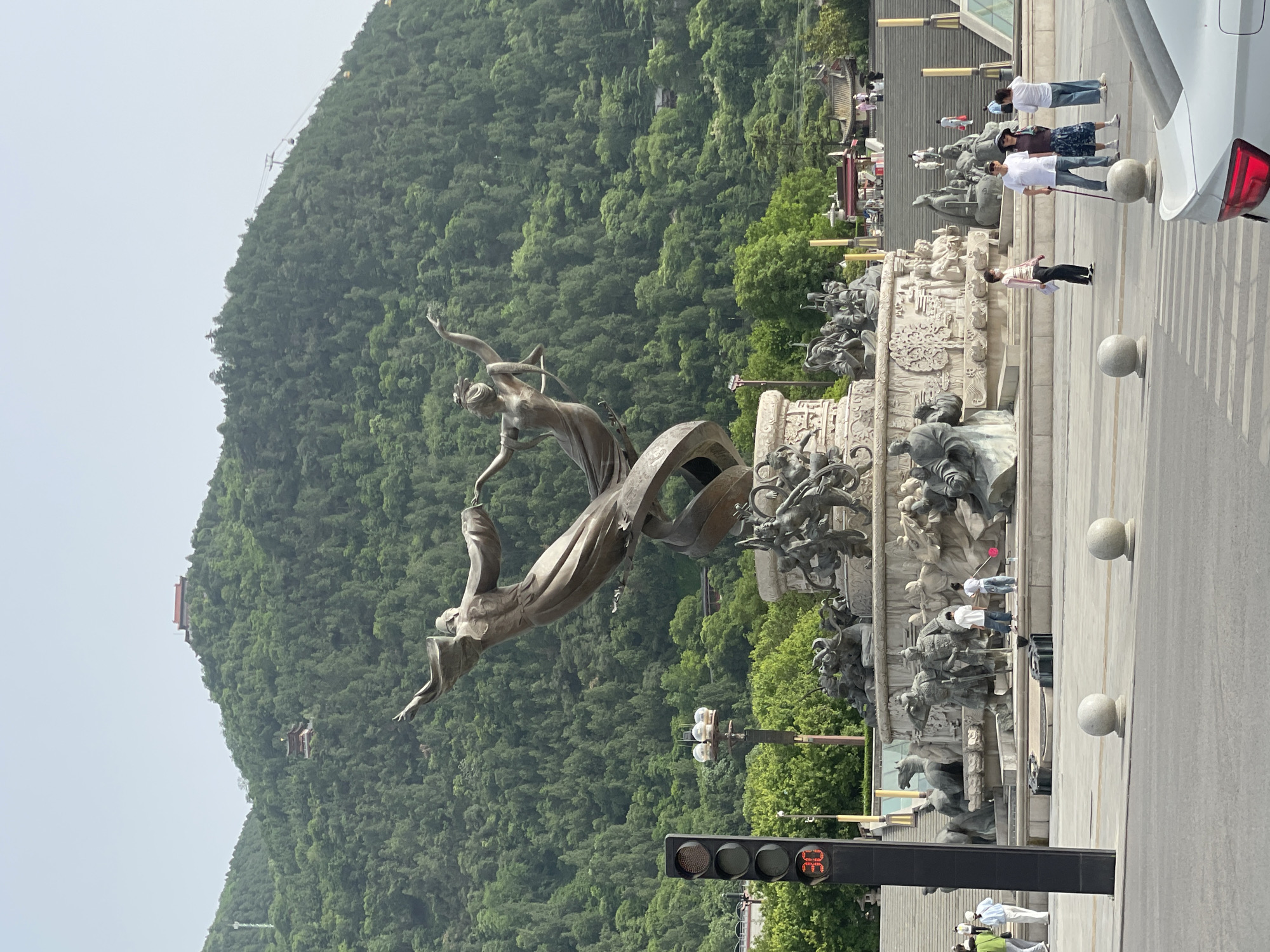 The image captures a large bronze sculpture in Lintong District, China, set against a backdrop of a densely wooded mountain. The sculpture depicts two figures in motion, with one figure reaching upwards. Below them, a complex arrangement of smaller sculptures is visible. The sculpture is located on a paved area adjacent to a road with a traffic light showing the number 36. People are seen walking along the edge of the paved area. A white car is partially visible on the right edge of the image. On the mountain slope, a building with a red roof can be seen. The sky is overcast, and the overall lighting is soft.