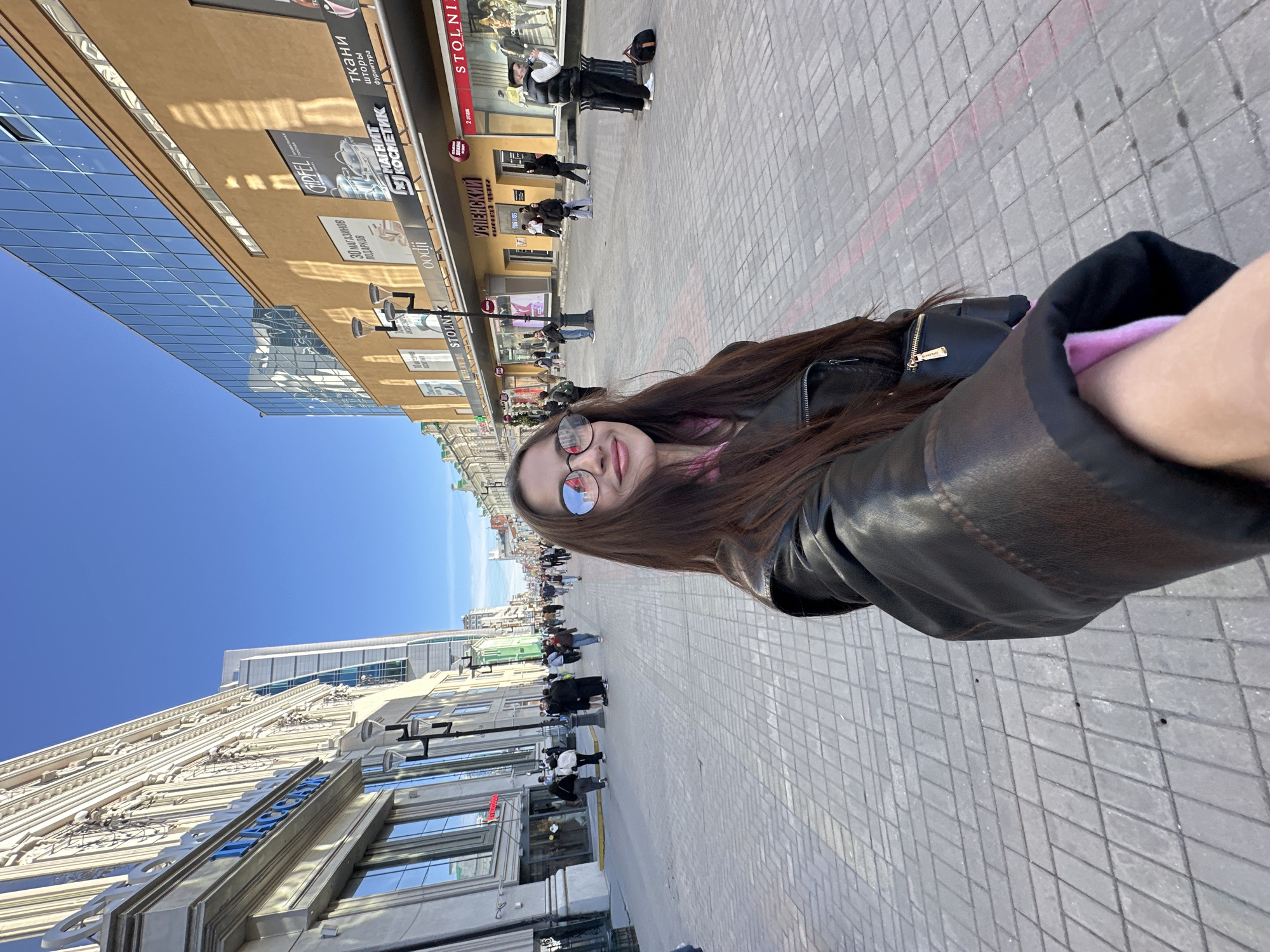The image is a selfie taken on Vaynera Street in Yekaterinburg, Russia. The subject is a young woman wearing glasses and a black leather jacket. She is standing on the sidewalk, which is made of grey paving stones. Buildings line both sides of the street, including a modern glass building on the left and buildings with storefronts displaying signage in Russian. "STOLNIK," "GIPFEL," "30 МАГАЗИНОВ ПОДАРКОВ," "oodji," "МАГНИТ КОСМЕТИК," "ткани шторы фурнитура," and "УСПЕНСКИЙ" are visible on the buildings. Pedestrians are seen walking along the street. The sky is bright blue.
