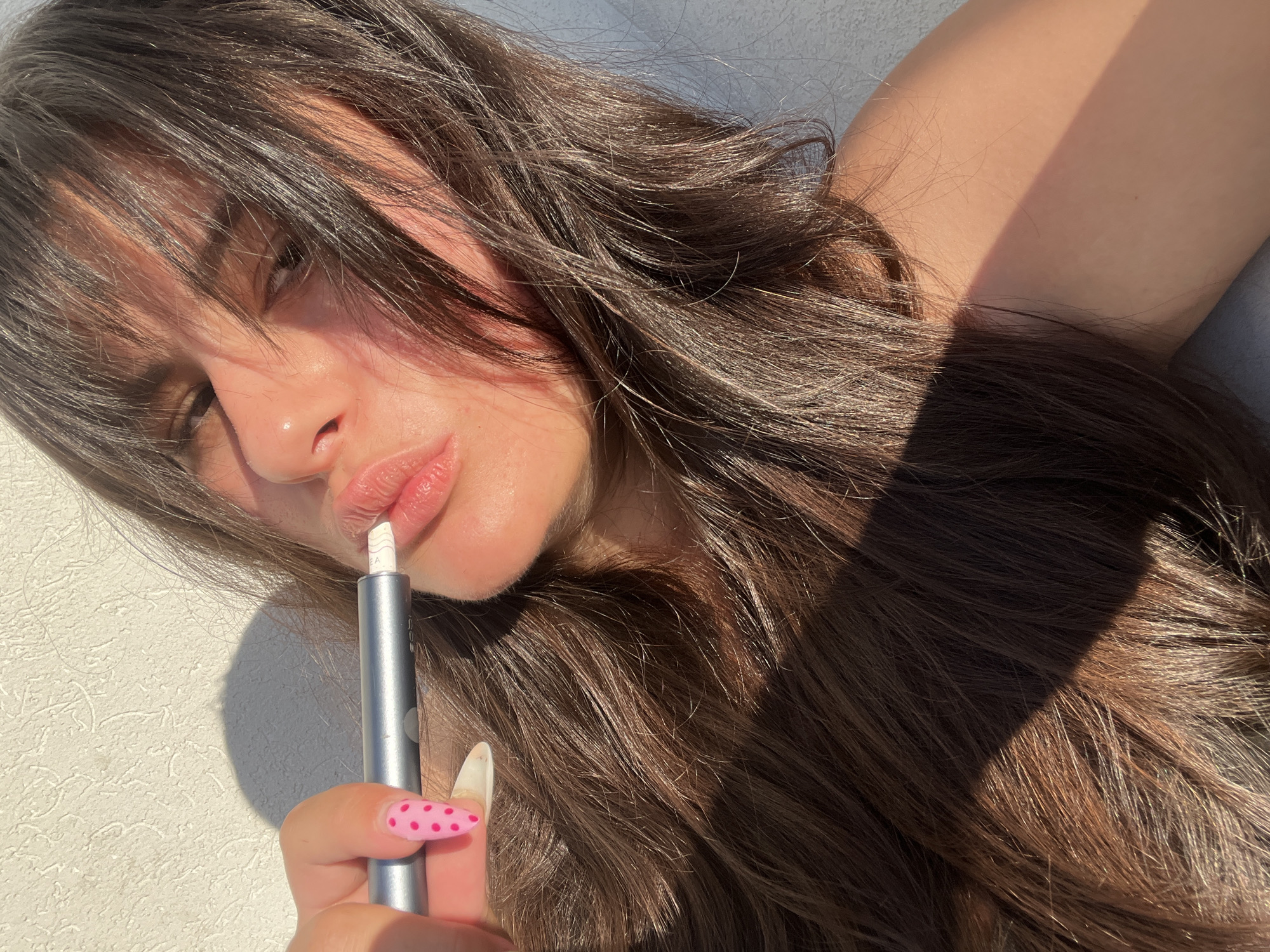 A close-up, high-angle shot shows a young woman with long, dark brown hair, partially covering her face, as she holds a silver pen-like object near her lips. She is wearing light-colored clothing and her nails are painted pink with white polka dots. The lighting suggests it is daytime, with sunlight casting a shadow across the scene. The background is a textured, light-colored wall, indicative of an outdoor setting. The image does not provide any specific details that can be used to determine a location in the city of Ashdod, Israel.