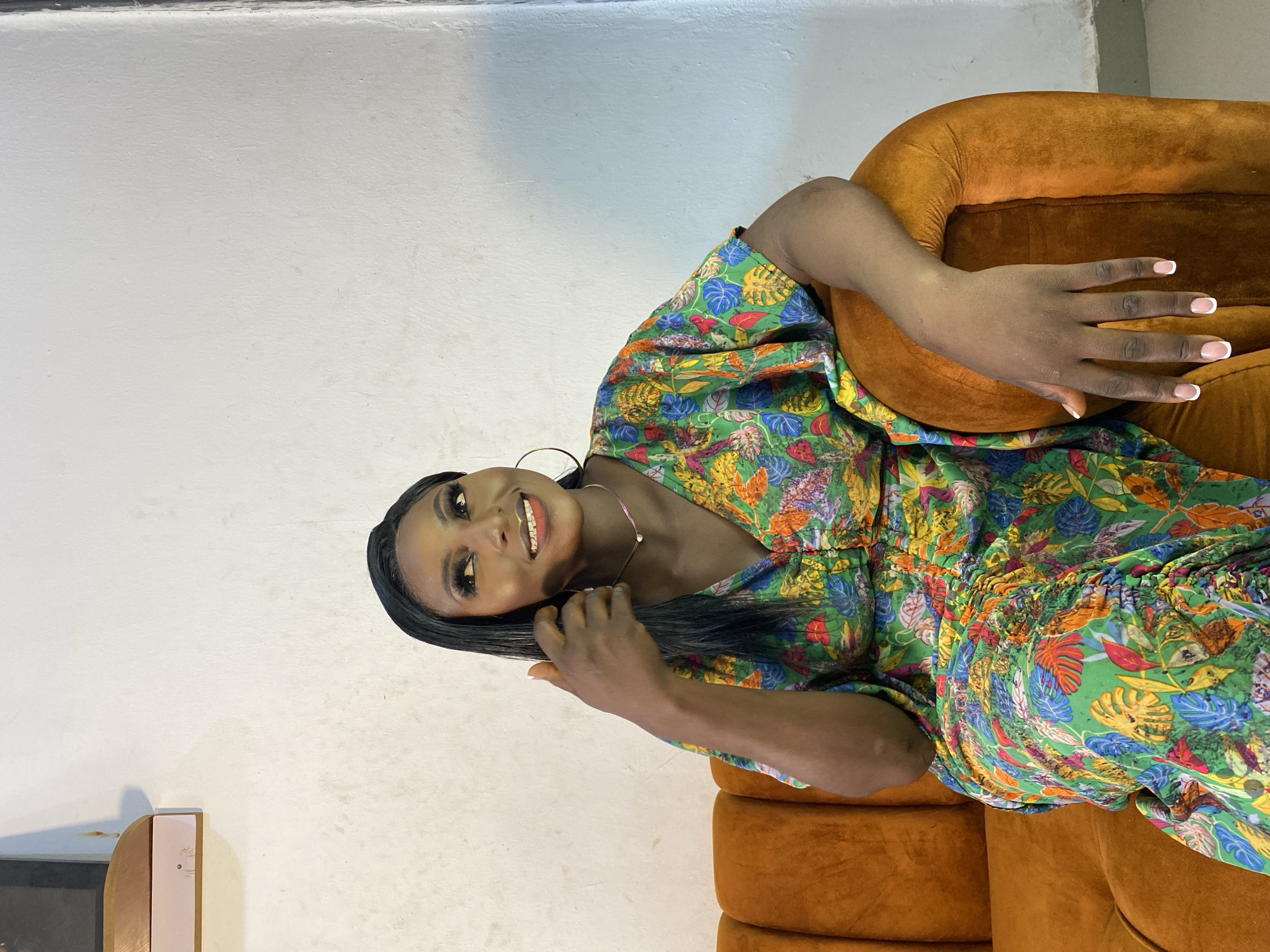 A woman with dark skin and long, straight black hair is sitting on an orange couch in front of a textured white wall. She is wearing a vibrant, multi-colored dress with a tropical leaf print. Her right hand is touching her hair, and her left arm is resting on the back of the couch. She is looking upwards and to her left with a subtle smile. She has on large hoop earrings and a delicate necklace. The background appears to be indoors, possibly in a residential setting.