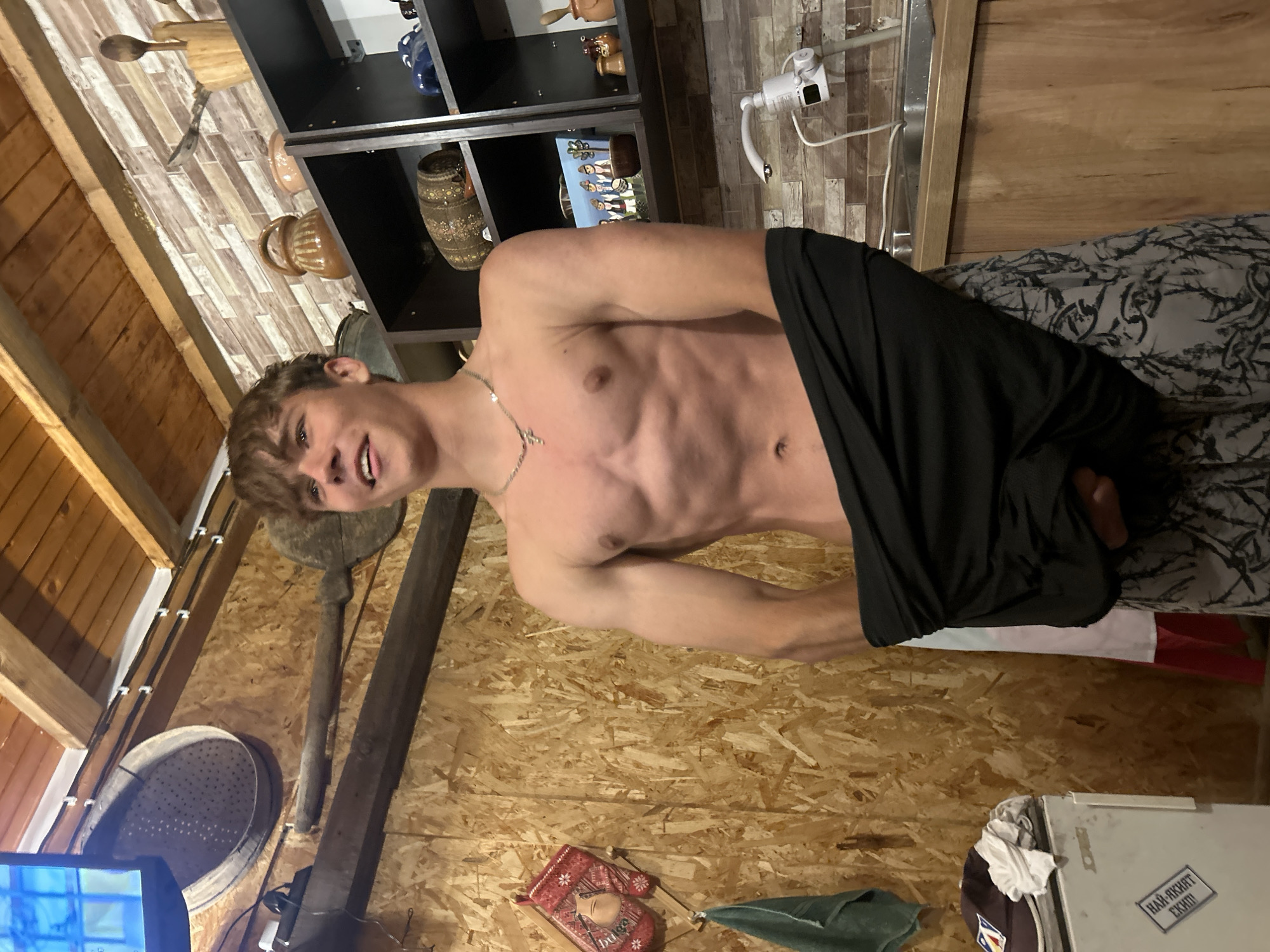 A young Caucasian male with light brown hair, wearing a silver cross necklace, stands shirtless in what appears to be a rustic kitchen area. He is wearing patterned shorts and is pulling a black shirt down. His abdomen is visible, showing well-defined musculature. The background features wooden walls and ceiling, shelves filled with various pottery and decorative items, a sink with a faucet, and a small refrigerator on the left side. A television screen is visible in the upper left corner. The lighting suggests an indoor setting, possibly a cabin or a traditional home.