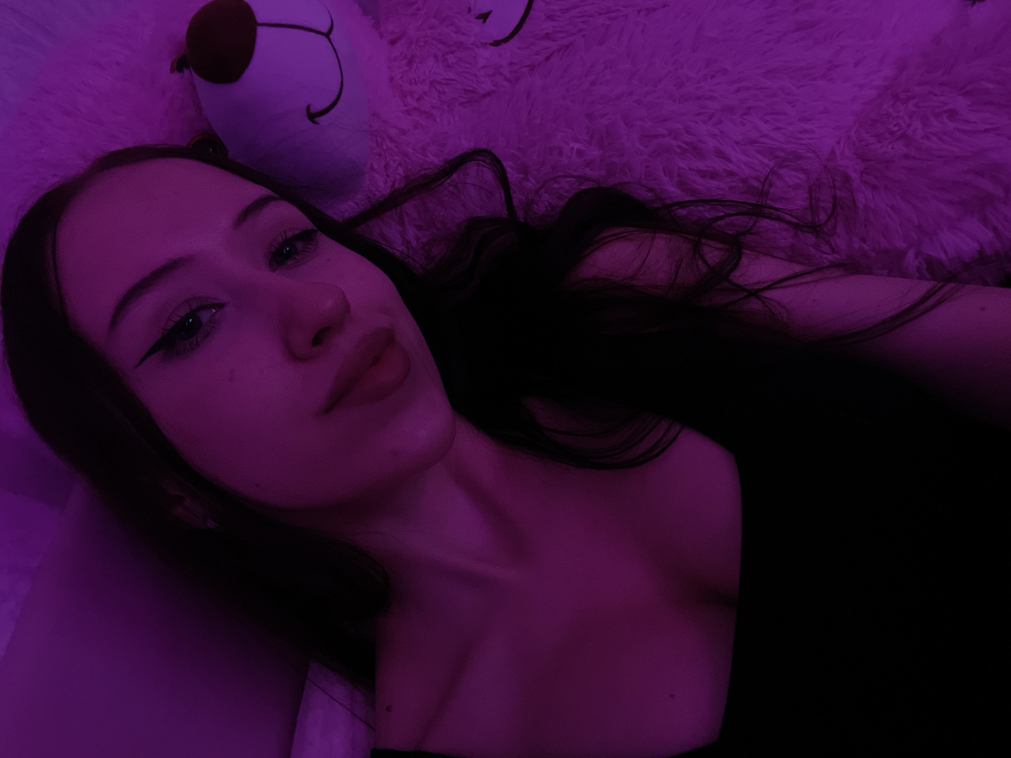 The image is a close-up, overhead selfie of a young woman with dark hair and winged eyeliner, illuminated by purple light. She is lying down next to a fluffy, light-colored stuffed animal, possibly a bear, which is visible on the right side of the frame. The woman is wearing a black top. The background is dark and appears to be indoors. The overall impression is of a casual, personal moment. The image does not contain any information that would indicate its location in Votkinsk, Russia.