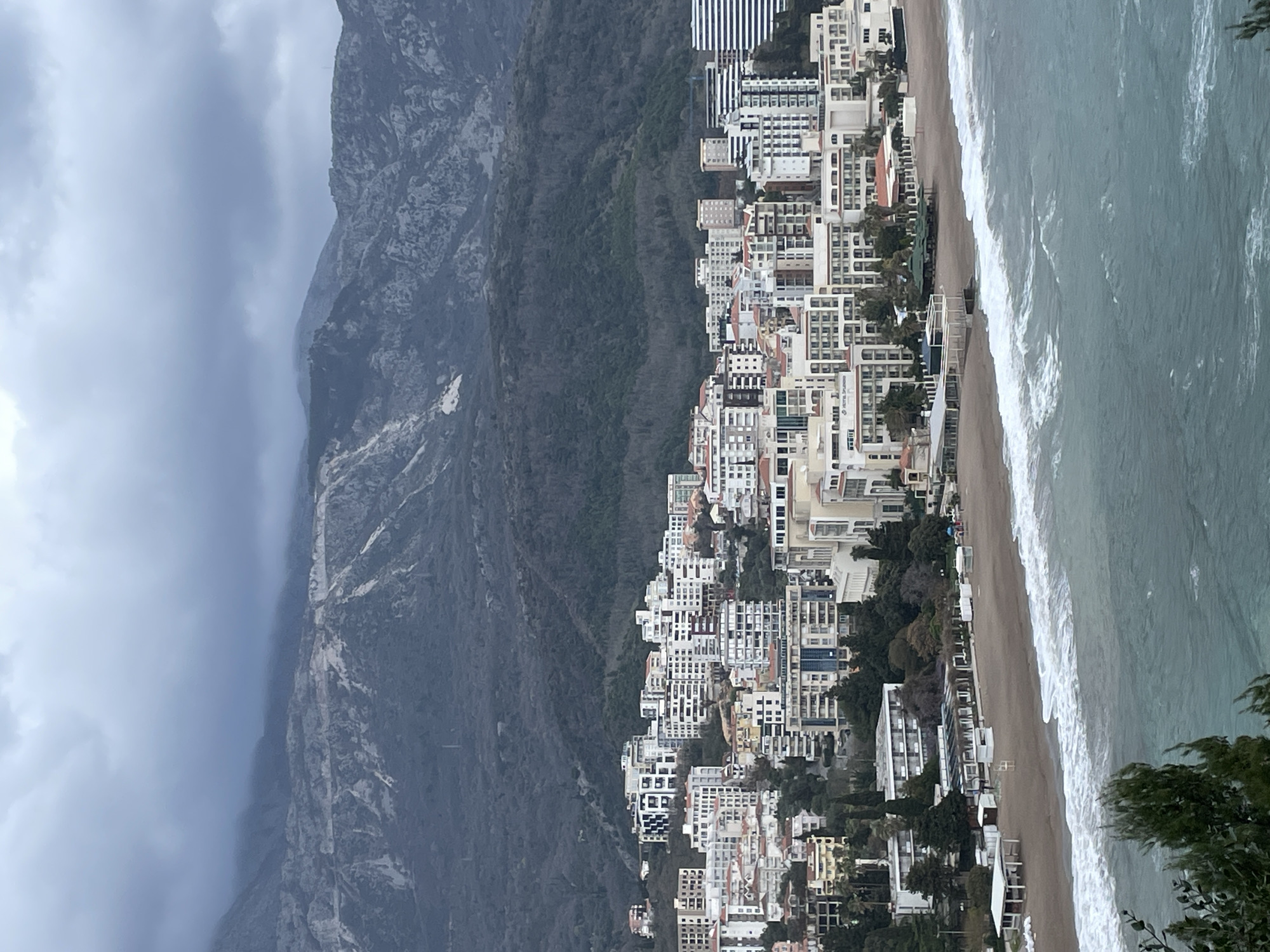 The image displays an elevated panoramic view of Boreti, Montenegro, under a heavily overcast sky with grey clouds. In the background, rugged, dark grey mountains rise prominently, featuring visible rock formations, sparse vegetation on higher slopes, and dense, dark green forests on their lower sections. The middle ground is occupied by a densely developed coastal town, consisting of numerous multi-story buildings, predominantly white or light-colored, with a mix of modern and traditional architectural styles. A large hotel complex, "Hotel Splendid," is clearly visible among these structures, situated closer to the beachfront. The foreground features a light brown sandy beach, which appears largely unoccupied. Several white, open-sided structures and covered facilities are present along the beach. The Adriatic Sea, characterized by a greenish-blue hue, borders the coastline, with moderate waves breaking into white foam along the shore. No individuals, specific actions, or interactions are discernible within the scene.
