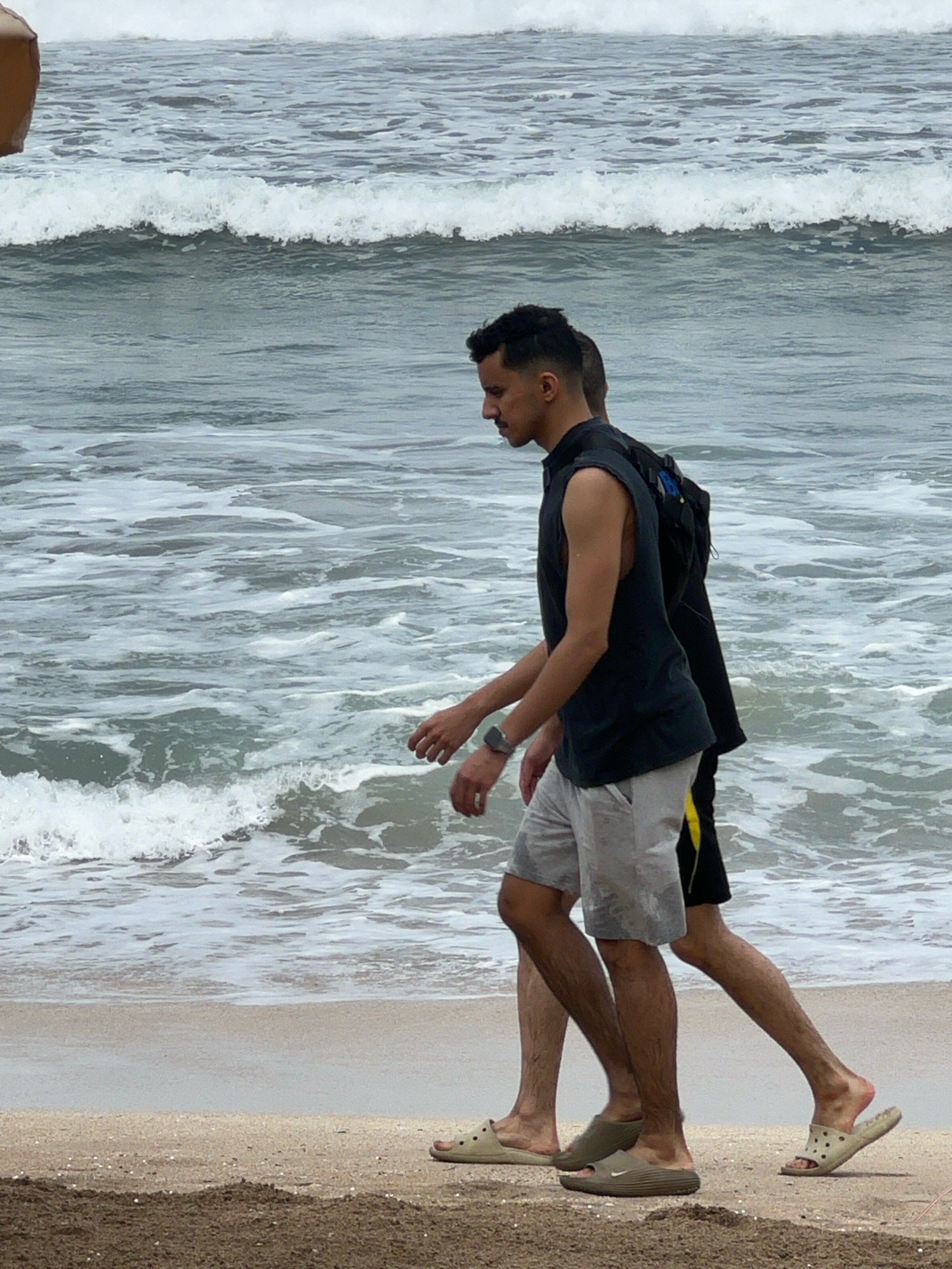 Two individuals walk side-by-side along the sandy shore of Kuta Beach, Bali, Indonesia. The man in the foreground is wearing a black sleeveless shirt and light grey shorts. He has a watch on his left wrist and is wearing light-colored slides. Behind him, another person, partially obscured, is wearing dark shorts with a yellow stripe and a backpack. The ocean, with moderate waves and white foam, stretches out behind them. The sky is overcast, contributing to the muted tones of the scene. The sand in the foreground is dark and appears damp.