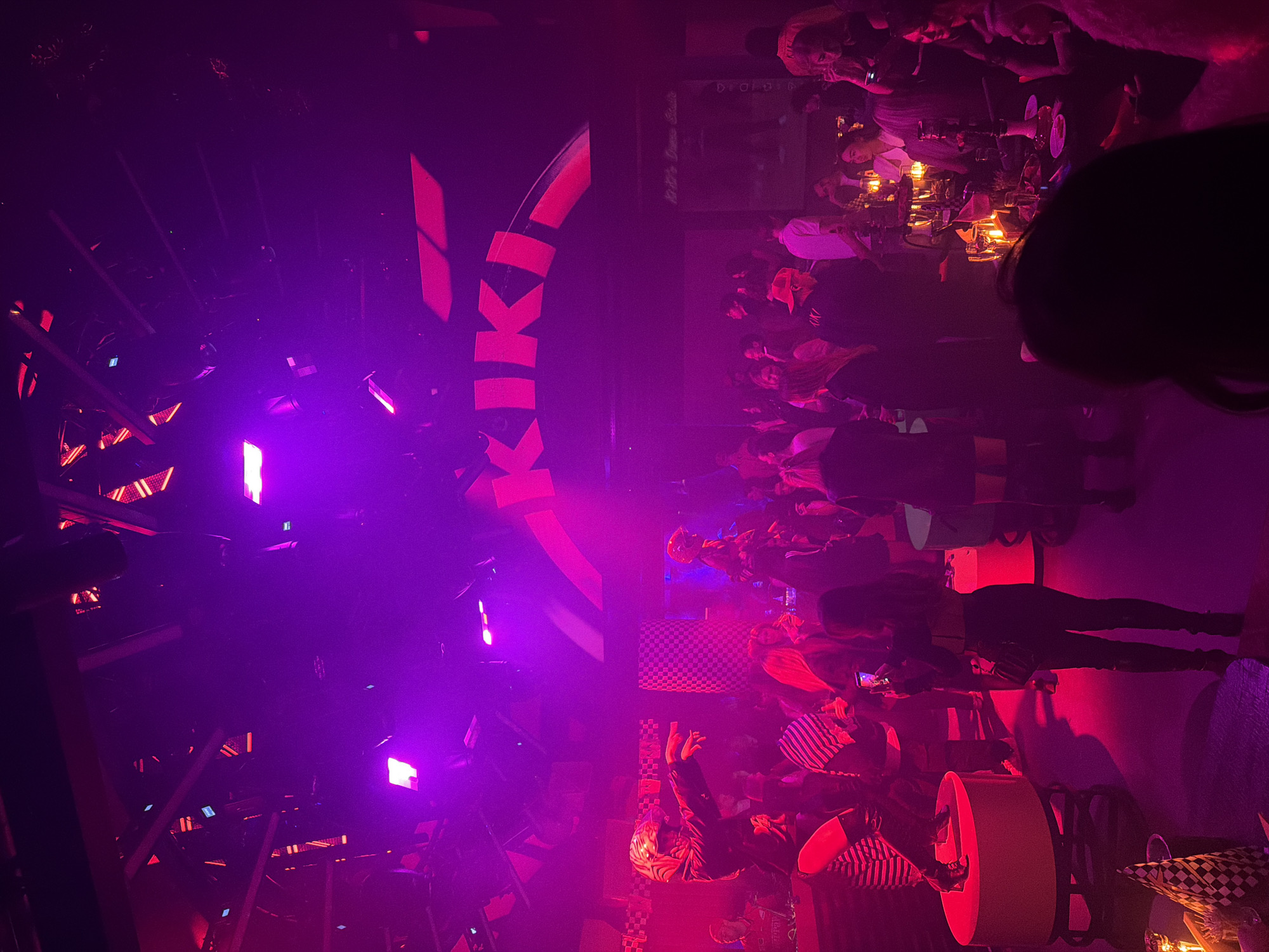 A dimly lit, crowded nightclub environment is depicted in the image, bathed in purple and red lighting. On the left, a large vertical sign displaying the word "KIKI" in stylized red letters is visible. Below the sign, lighting fixtures and structural elements of the club's interior are visible. The right side of the image reveals a group of people, some seated at tables with illuminated drinks. Individuals are standing and interacting, some holding phones. A checkerboard pattern is present on some of the seating and decor. The overall atmosphere suggests a lively nightlife scene. It is not possible to confirm the city location as Moscow, Russia from the given visual information.