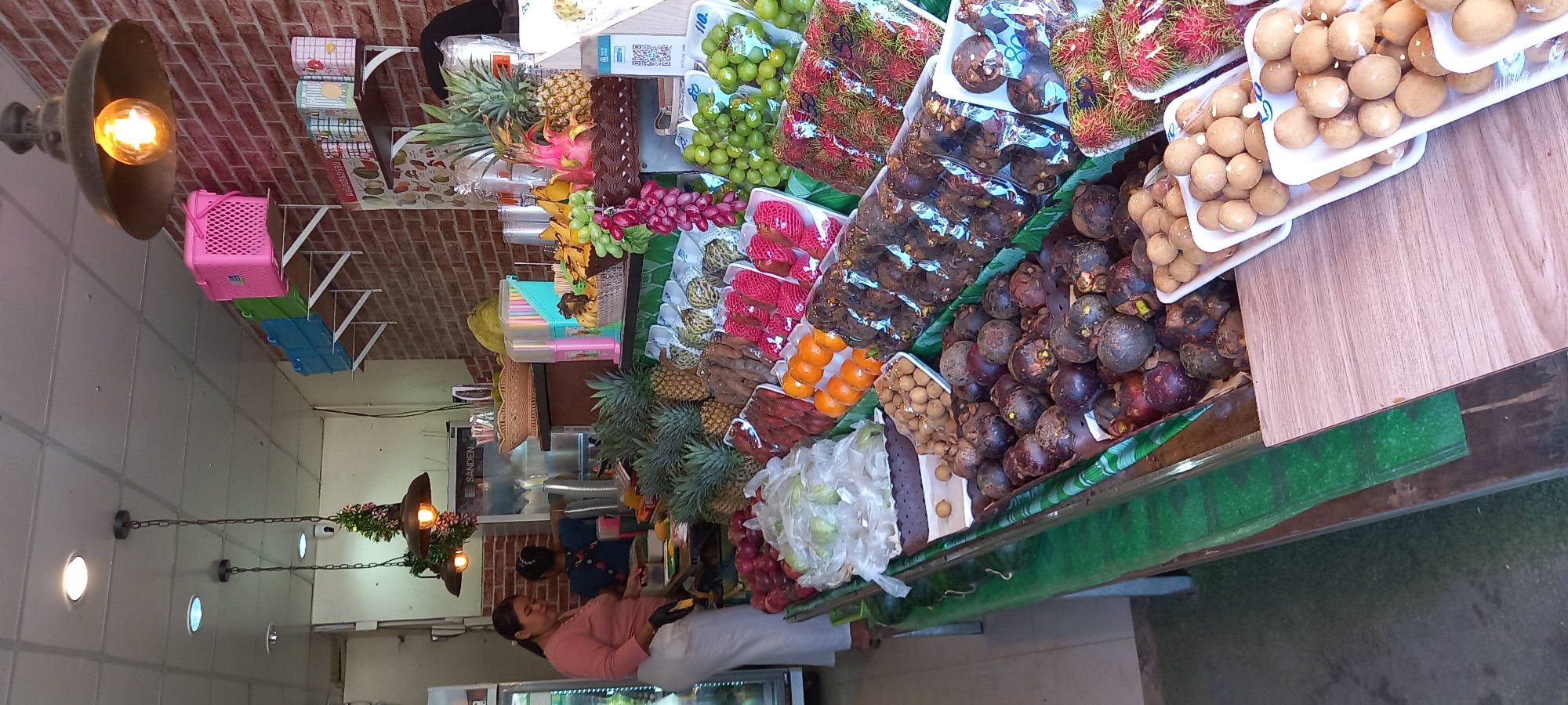 This is an image of a fruit stand in Ban Ao Nang, Thailand. The stand displays a variety of tropical fruits, including pineapples, grapes, bananas, dragon fruit, rambutans, mangosteens, and longans. A woman stands behind the stand, seemingly tending to the fruit. The stand is decorated with green trim. There are lights hanging above and to the side. Shelves with miscellaneous items are visible in the background, as is a brick wall.