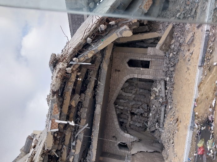 A severely damaged building lies in ruins, its structure fractured and crumbling. Rebar and concrete debris are scattered throughout the scene, indicating significant structural collapse. Arched architectural elements are still visible, suggesting a distinctive design now marred by destruction. Rubble fills the remaining open spaces, contrasting with the relatively clear ground beside the building. Debris and trash are also seen on the street, highlighting the aftermath of a devastating event.