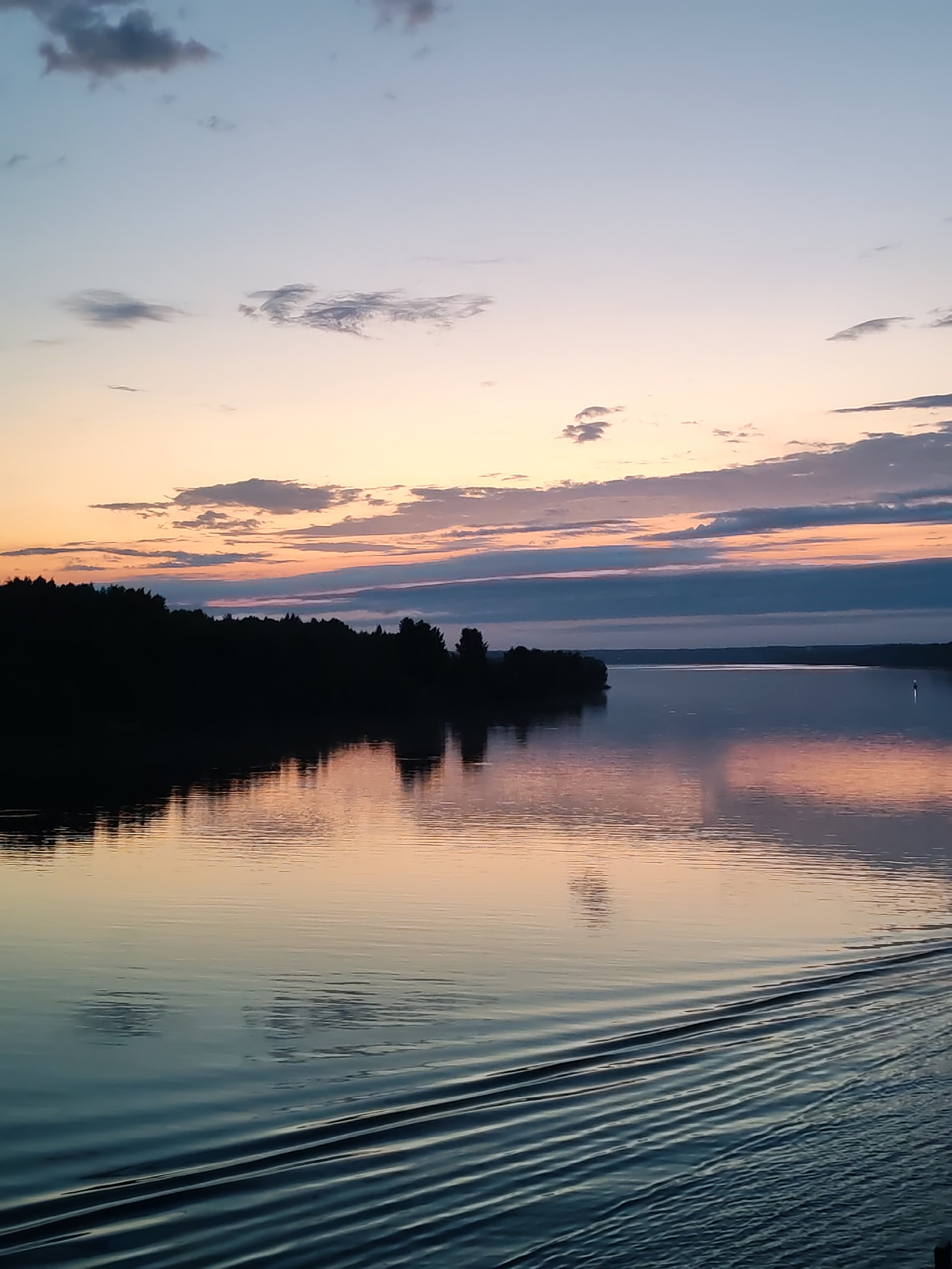 The image depicts a body of water reflecting a colorful sunset sky. The sky transitions from pale blue at the top to shades of orange and pink near the horizon. Dark clouds are scattered throughout the sky. A dark treeline is visible on the left side of the image, likely representing the shoreline. The water is calm with ripples caused by a boat or other watercraft in the lower right. A small white object is visible in the distance. The location is reported to be Baskachi, Russia.