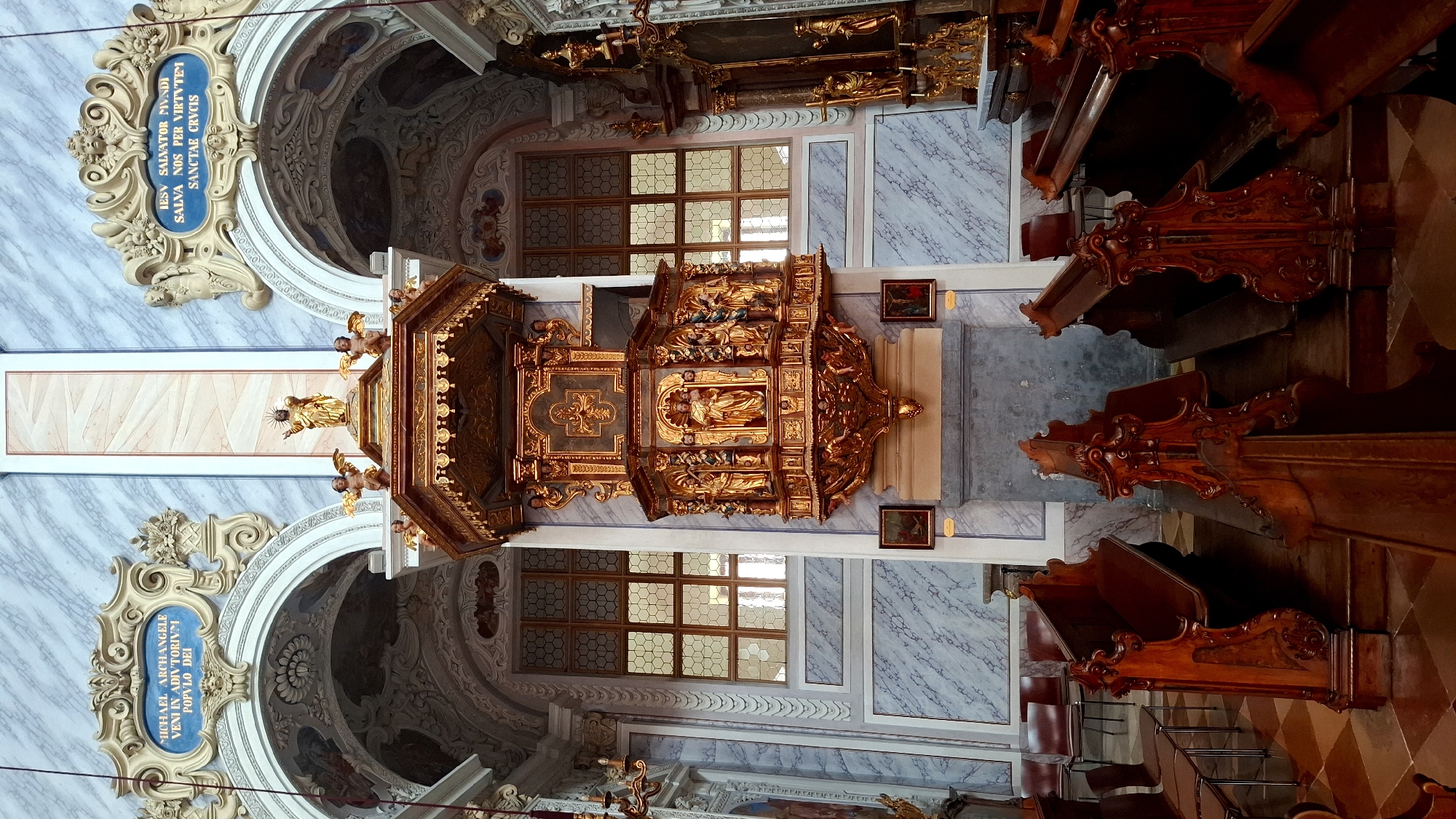 The image depicts the interior of a Baroque-style church, likely Stift Göttweig Abbey in Furth bei Göttweig, Austria, specifically focusing on the ornate pulpit and surrounding pews. The pulpit is a grand, gilded structure with multiple tiers, adorned with sculptures of religious figures and intricate detailing. Above the pulpit, a golden statue of Christ stands prominently. Flanking the pulpit are arched windows with intricate lattice work, and the walls feature a marble-like pattern. In the foreground, dark wooden pews with elaborately carved ends are arranged in rows, facing the pulpit. The floor has a checkered pattern of brown and beige tiles. The overall impression is one of opulence and religious grandeur, characteristic of Baroque church architecture.
