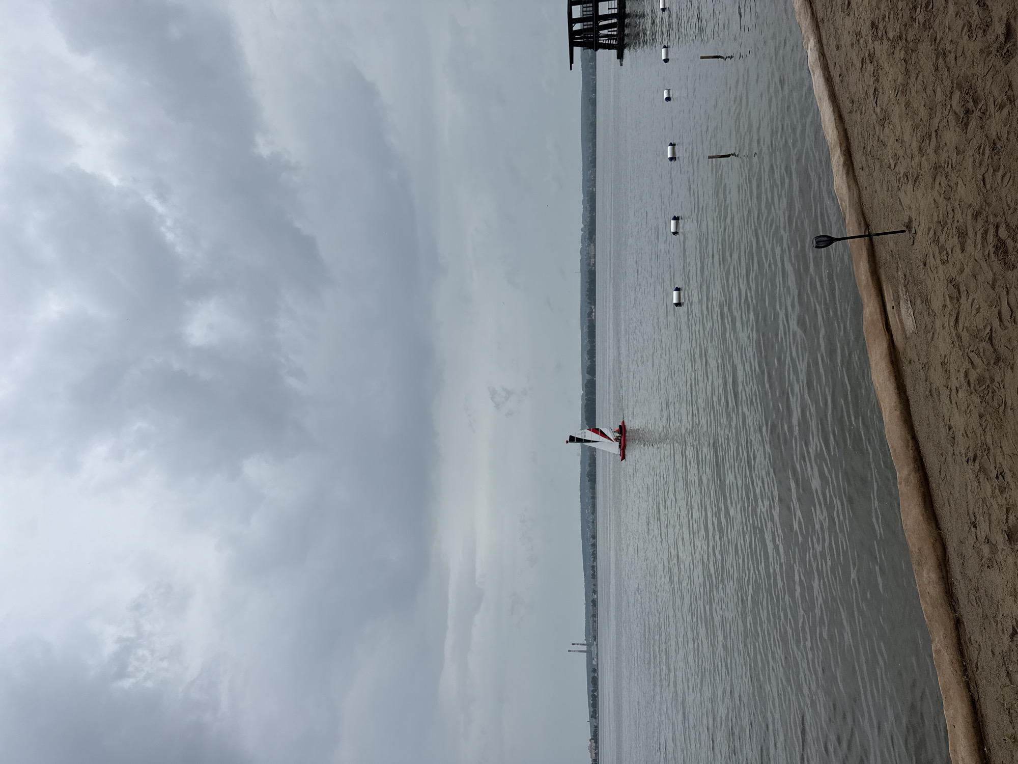 A sailboat with red and white sails navigates a calm body of water under a cloudy sky. In the distance, a shoreline is visible with trees and industrial structures. On the right side of the image, a dark wooden pier or dock extends into the water. The foreground features a sandy beach with a large log and a decorative solar-powered torch. Several white buoys are lined up in the water. This scene is likely located on the Votkinsk Reservoir in Votkinsk, Russia, given the characteristic industrial silhouette and the presence of a large body of water.
