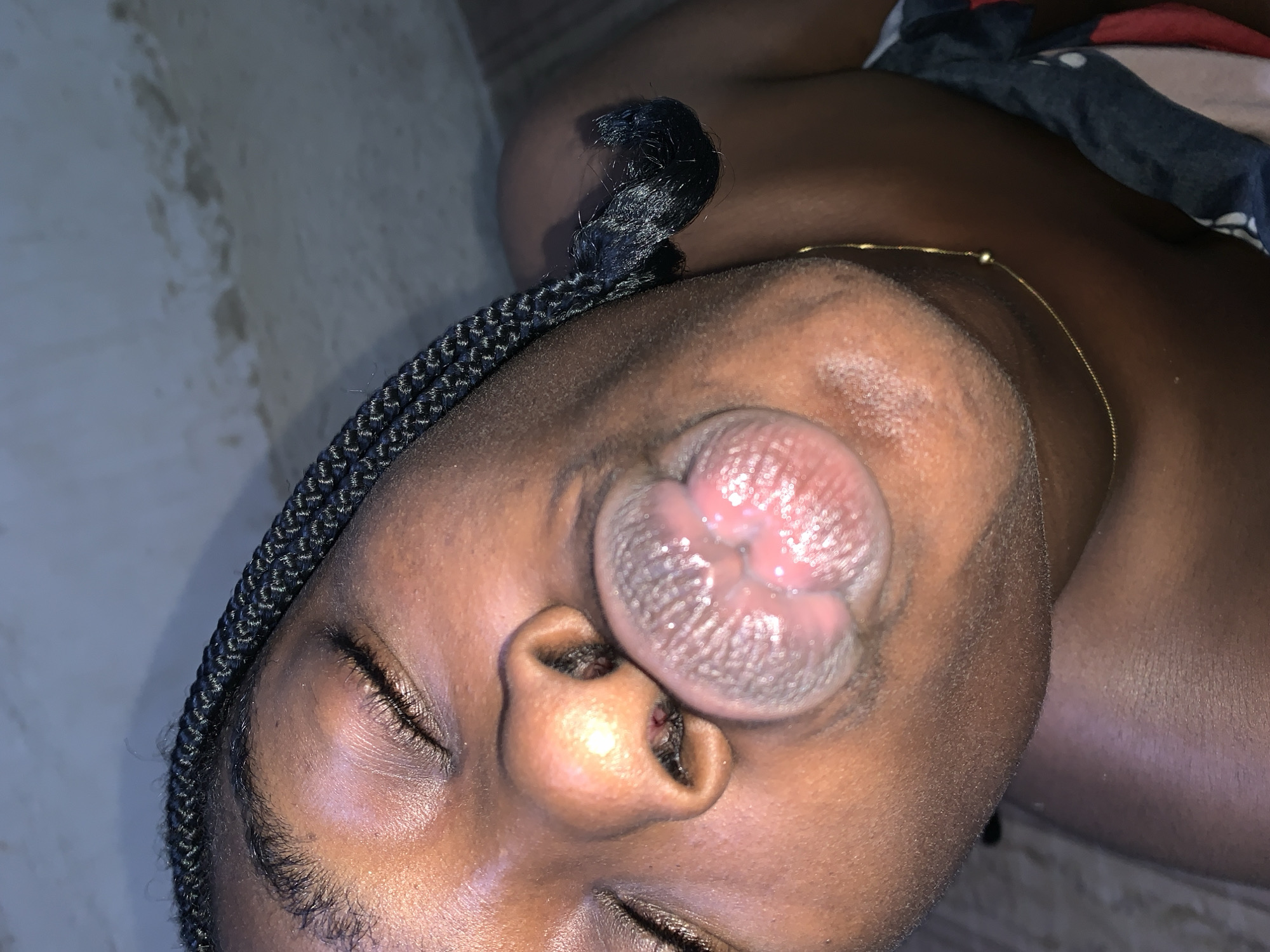 The image shows a close-up of a person's face, with their eyes closed and their lips pursed. The person has dark skin and braided hair. They are wearing a thin gold chain around their neck. The background appears to be a textured wall. The scene is indoors, likely in a domestic setting. There is no discernible location in the city of Rafin Par, Nigeria.