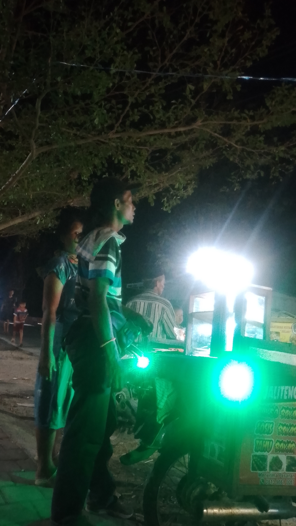 The image depicts an outdoor scene at night, possibly in Karanglangu, Indonesia. In the foreground, a person wearing a striped shirt and a dark cap stands facing right, illuminated by a bright green light. Behind them, another person in a blue shirt and dark shorts stands. To the right, a food cart with a bright light source is visible, with signage that reads "ALITENG" and lists items like "SOSIS BAKAR" and "TAHU BAKAR," indicating it is a grill stall. An older man in a striped shirt and a cap is seated behind the cart. The background shows trees and darkness, suggesting a rural or less developed urban setting. Several other individuals, including children, are faintly visible in the background, some appearing to be customers. The overall atmosphere appears to be that of a nighttime street food vendor operating in a community setting.