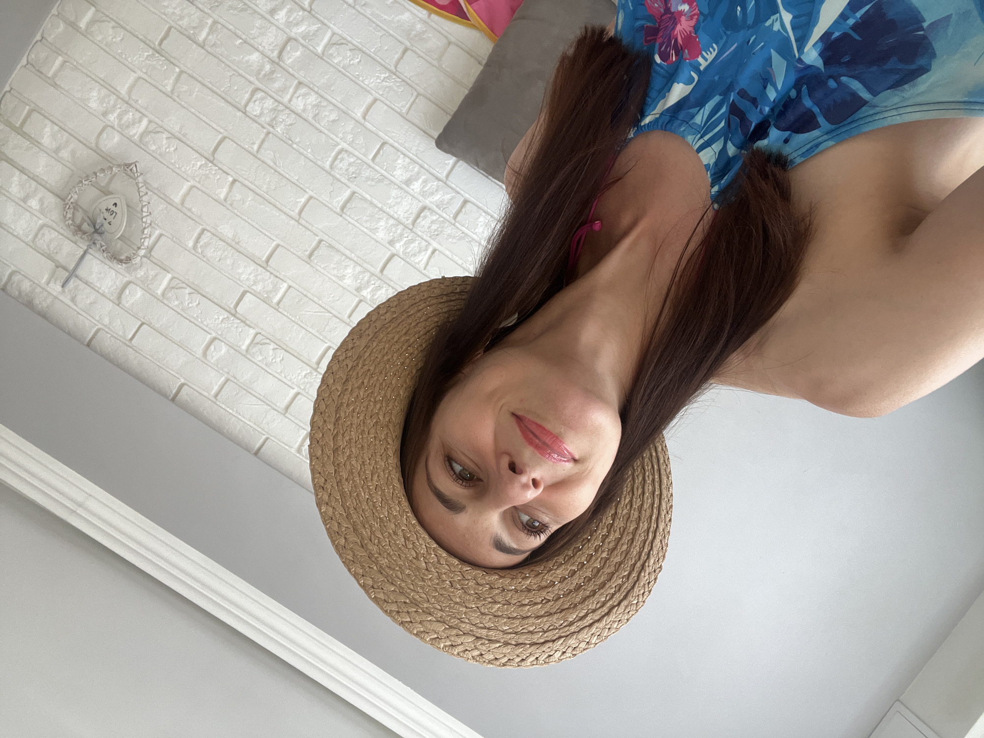 This media file shows a woman wearing a straw hat and a blue and floral patterned top. She has long, dark brown hair and is wearing makeup. The woman is looking towards the camera with a slight smile. The background is a white brick wall and a grey ceiling. There is also a hint of a grey pillow and a pink item in the background. The image appears to be taken indoors. There is no visible text. The lighting suggests it is daytime. The woman's attire and the hat suggest a casual, possibly summery mood.