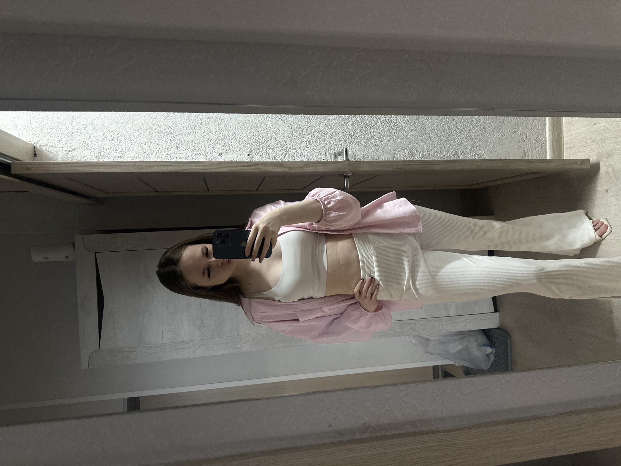 A young woman is captured in a mirror selfie, standing upright and posing indoors. She holds a dark-colored smartphone with her right hand, angled slightly away from her face, while her left hand rests near her hip. She has long, dark hair and a neutral expression, looking at her phone.
She is dressed in a casual yet stylish outfit, consisting of a white crop top, white ribbed flared pants, and a light pink oversized button-up shirt worn open over the top. Her sleeves are slightly puffed at the shoulders and gathered at the wrists. She is wearing light-colored, open-toed sandals or slides on her feet.
The background, visible through the mirror's reflection, depicts what appears to be a home interior. The walls are a light color, possibly white or off-white, with a visible texture. Light-colored wooden furniture or paneling is present on either side of her and forming a shelf or counter behind her. The floor is covered with light wood-colored planks. In the lower right corner of the reflection, a black or dark grey object resembling a bathroom scale is visible on the floor, next to a white plastic bag, partially tucked under a shelf.
The primary activity is the woman taking a mirror selfie to showcase her outfit. The setting suggests a casual moment within a personal space, likely a bedroom or dressing area. No specific time of day or weather conditions are discernible, and there is no visible text in the image.