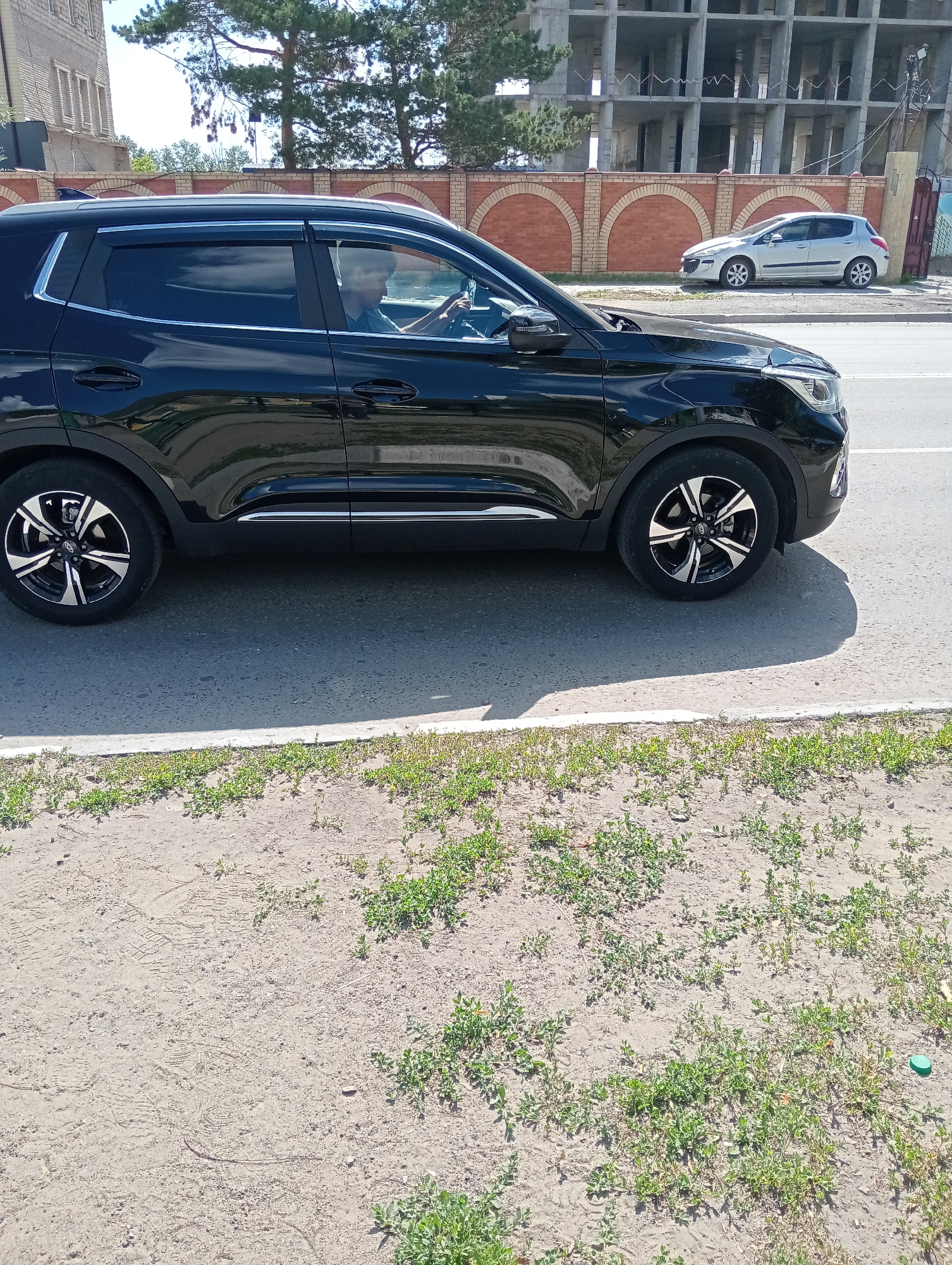 A black SUV is parked on the side of a road, with its front facing towards the right of the frame. A person is visible in the driver's seat, looking forward. In the background, there is a construction site with a partially built structure. To the right of the SUV, a silver hatchback is parked. The ground in front of the SUV is covered with dirt and some green vegetation. A tree with green foliage is visible behind a brick wall. The sky is blue and appears to be clear.