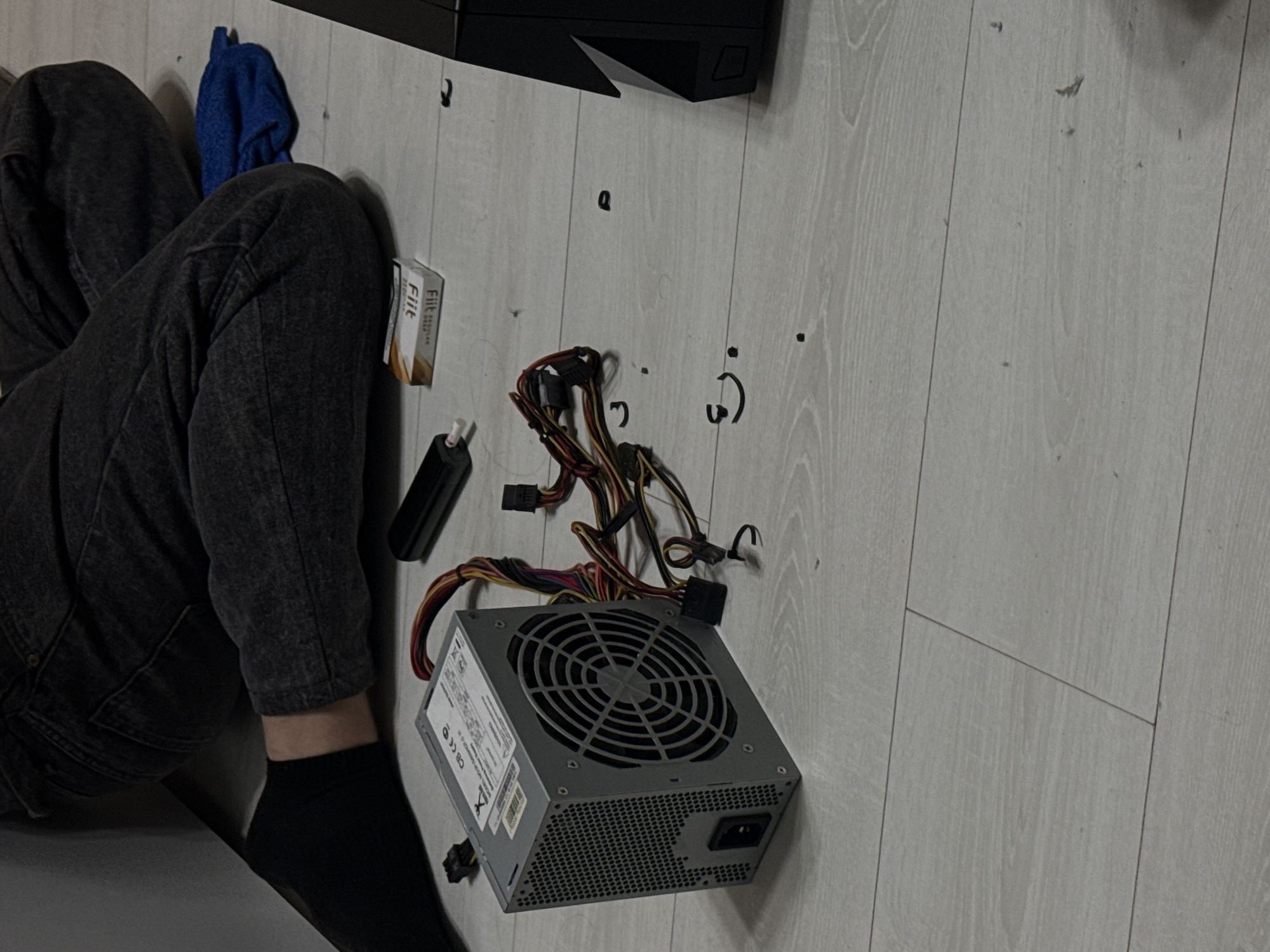 The image depicts a low-angle shot on a light-colored, wood-grained floor. A person is partially visible, wearing dark grey jeans and a black sock. Next to the person are several objects related to computer hardware. A gray computer power supply unit (PSU) is prominently displayed with various colored wires and connectors extending from it. Near the PSU is a black object and a small box of "Fiit" brand items. A crumpled blue cloth is near the top left, partially obscured by the jeans. The lighting appears to be indoor and overhead.