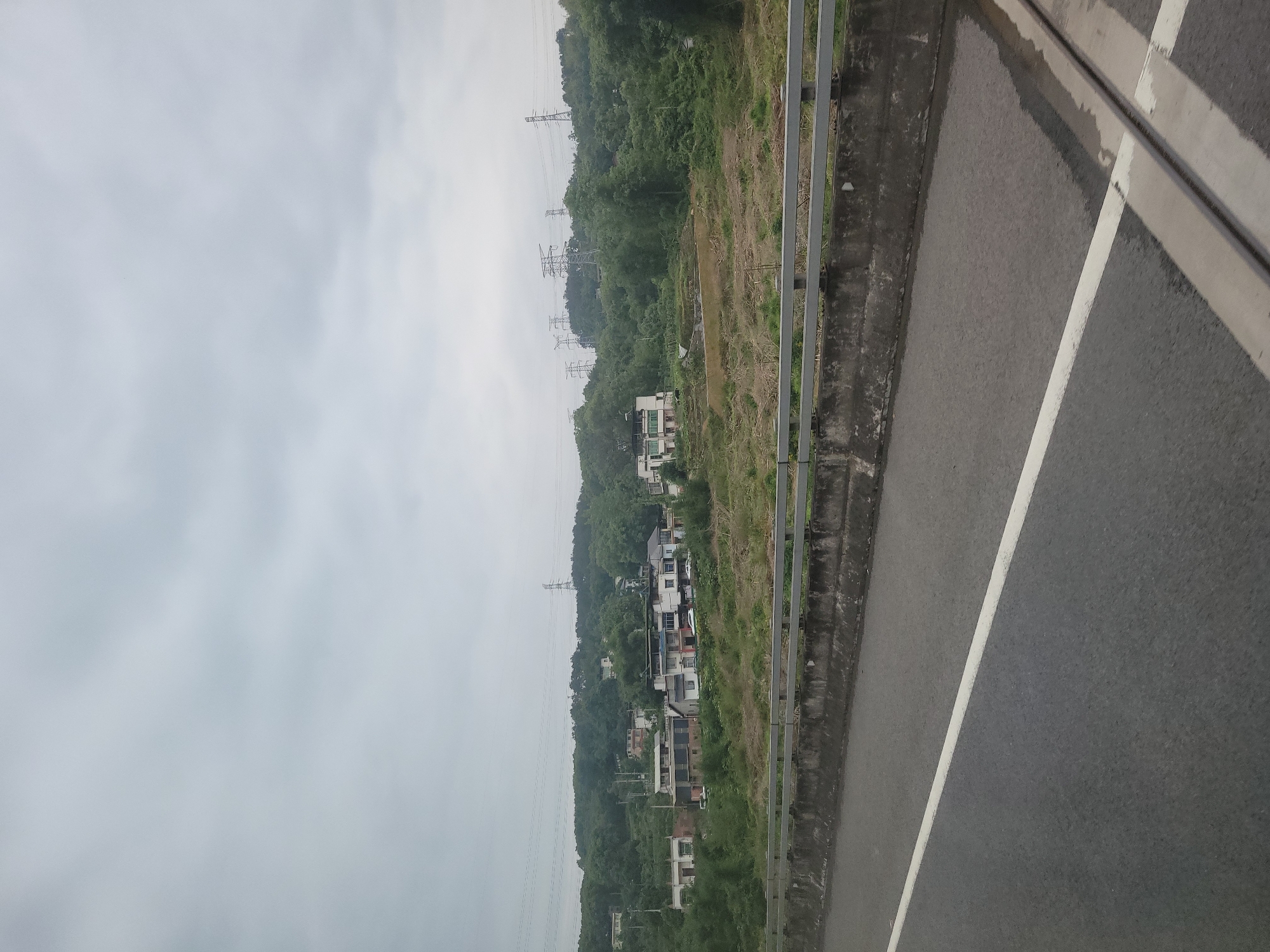 The image depicts a roadside scene in Beibei, China. A gray sky dominates the upper portion of the frame. A line of utility poles ascends a green hillside with interspersed structures, including residential buildings. A gray metal guardrail and concrete barrier run along the roadside. The foreground consists of a gray asphalt road with a white lane marking.