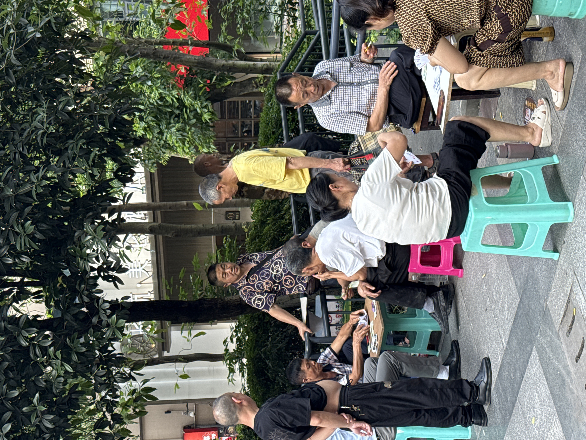 This outdoor scene, likely captured in the Yuzhong District of China, depicts a vibrant gathering of predominantly older adults engaged in leisure activities. The setting is a paved area, possibly a park or public square, shaded by dense green trees. Buildings with red accents are visible in the background, peeking through the foliage.
At the heart of the activity, several individuals are seated around small, low wooden tables, using colorful plastic stools (teal, pink, blue). The primary activity appears to be playing cards, with participants holding hands of cards and observing the game intently. One woman, wearing a brown patterned dress, holds cards and has a lit cigarette, suggesting a relaxed, social atmosphere. Other men are seen participating, leaning over tables, or standing nearby, observing the card games. Their attire is casual, consisting of t-shirts, shirts, and trousers, suitable for a daytime outdoor setting.
The overall mood is one of engagement and social interaction. People are focused on the games, some standing and watching, others actively playing. The bright daylight indicates it's daytime, and the presence of outdoor activities suggests pleasant weather. Visible objects include playing cards, various plastic stools, small tables, and thermos bottles, common items for outdoor socializing in China. No clearly legible text is discernible in the image.