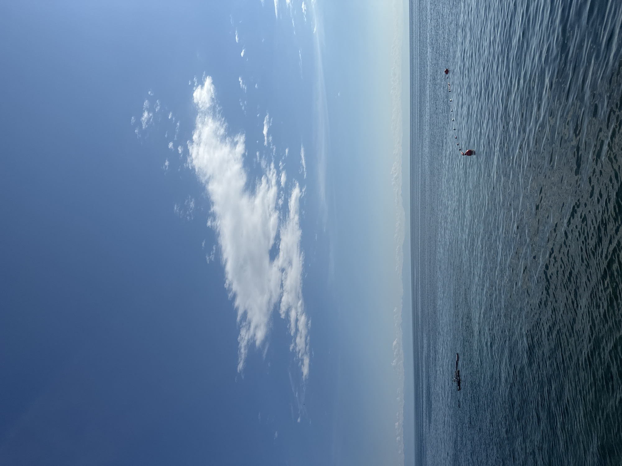 The image depicts a serene seascape with a clear blue sky filled with scattered white clouds. The calm blue water stretches to the horizon where a distant mountain range is faintly visible. In the foreground, a line of buoys marks a section of the water, with two larger red buoys more prominently placed. To the left, a small, dark boat with a structure on it is visible on the water. The overall scene suggests a peaceful day on the water, potentially near the shore of Lake Baikal in Listvyanka, Russia, given the presence of buoys and the mountainous backdrop characteristic of the region.
