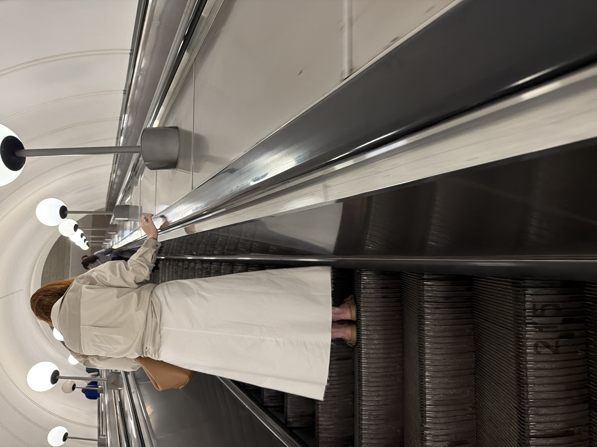 The image depicts the interior of a Moscow Metro station, specifically on an escalator. The perspective is from behind a woman with auburn hair, wearing a light-colored oversized shirt and a long white skirt. She is holding a tan tote bag over her left shoulder and is walking up the escalator. Her feet are visible, wearing light brown sandals.
The escalator's metal steps are dark and ribbed, showing signs of wear. To the right of the escalator is a polished metal handrail. The ceiling of the station is arched and white, illuminated by rows of spherical, globe-like lights mounted on grey poles. In the distance, other passengers can be seen on the escalator, and some are walking in the opposite direction. The overall impression is that of a typical, albeit well-lit, Moscow Metro station environment.