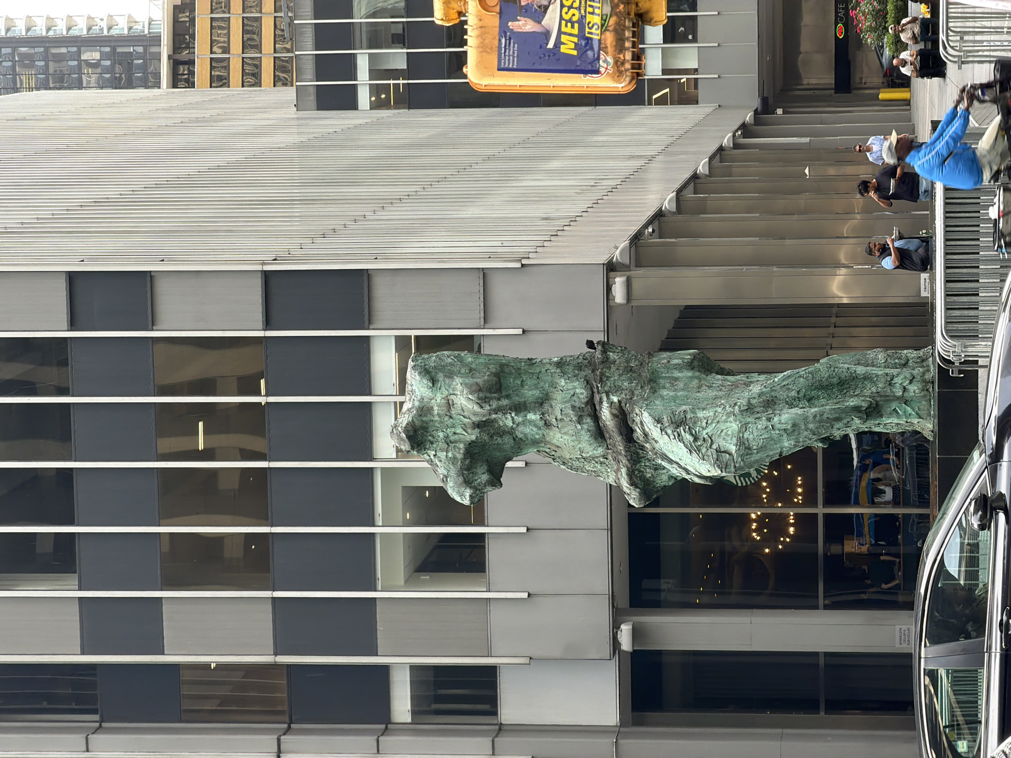 This image depicts a street scene in New York City, United States of America, likely Manhattan given the architecture and signage. In the foreground, a large, abstract, green-patinated bronze sculpture of intertwined figures stands prominently. Behind the sculpture, a modern skyscraper with a facade of alternating dark and light panels and vertical silver fins dominates the background.
Several people are visible. To the right of the sculpture, three men are standing and eating from bowls. Further to the right, a man wearing a blue jacket is riding a bicycle with a cargo carrier. A yellow traffic signal, featuring an advertisement for "MESS IS HERE," is mounted on a pole to the right of the sculpture. In the mid-ground, a black car is parked on the street. Through the large glass windows of the building behind the sculpture, interior lights and reflections of chandeliers are visible. The overall environment suggests an urban setting with a mix of public art and commercial architecture.