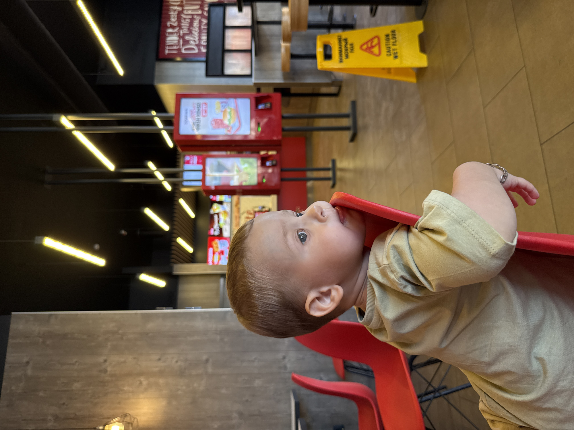 The image presents a candid view of a young child seated in a commercial dining establishment in Rusanovo, Russia. The child, appearing to be a toddler, is positioned in the foreground, seated in a red plastic chair and facing right in profile. The child has light brown hair, blue eyes, and is wearing a light olive-green short-sleeved top. A small, clear liquid droplet is visible beneath the child's right nostril. The child's right arm, adorned with a silver chain bracelet, rests on the red chair's backrest or arm.
The background reveals the interior of a modern restaurant or cafeteria. To the left, a vertical wall panel featuring a light wood grain texture is visible, along with a caged pendant light fixture hanging from a dark ceiling. Another empty red chair is partially visible to the left of the child. The ceiling is black and features multiple linear light fixtures emitting a warm yellow glow, arranged at various angles.
In the midground and background to the right, several red-framed self-service kiosks with digital screens display images of food items, including burgers and beverages. These kiosks stand upright on a light brown tiled floor. Adjacent to the kiosks, a yellow A-frame "CAUTION WET FLOOR" sign is prominent, displaying text in both Russian ("ВНИМАНИЕ! МОКРЫЙ ПОЛ") and English. Further into the background, several tall tables with round wooden tops and black bases are visible, along with a wall-mounted sign containing partially visible English text, including phrases like "TENDER Zesty & Tangy BUT DELICIOUS." The overall lighting is bright and even.