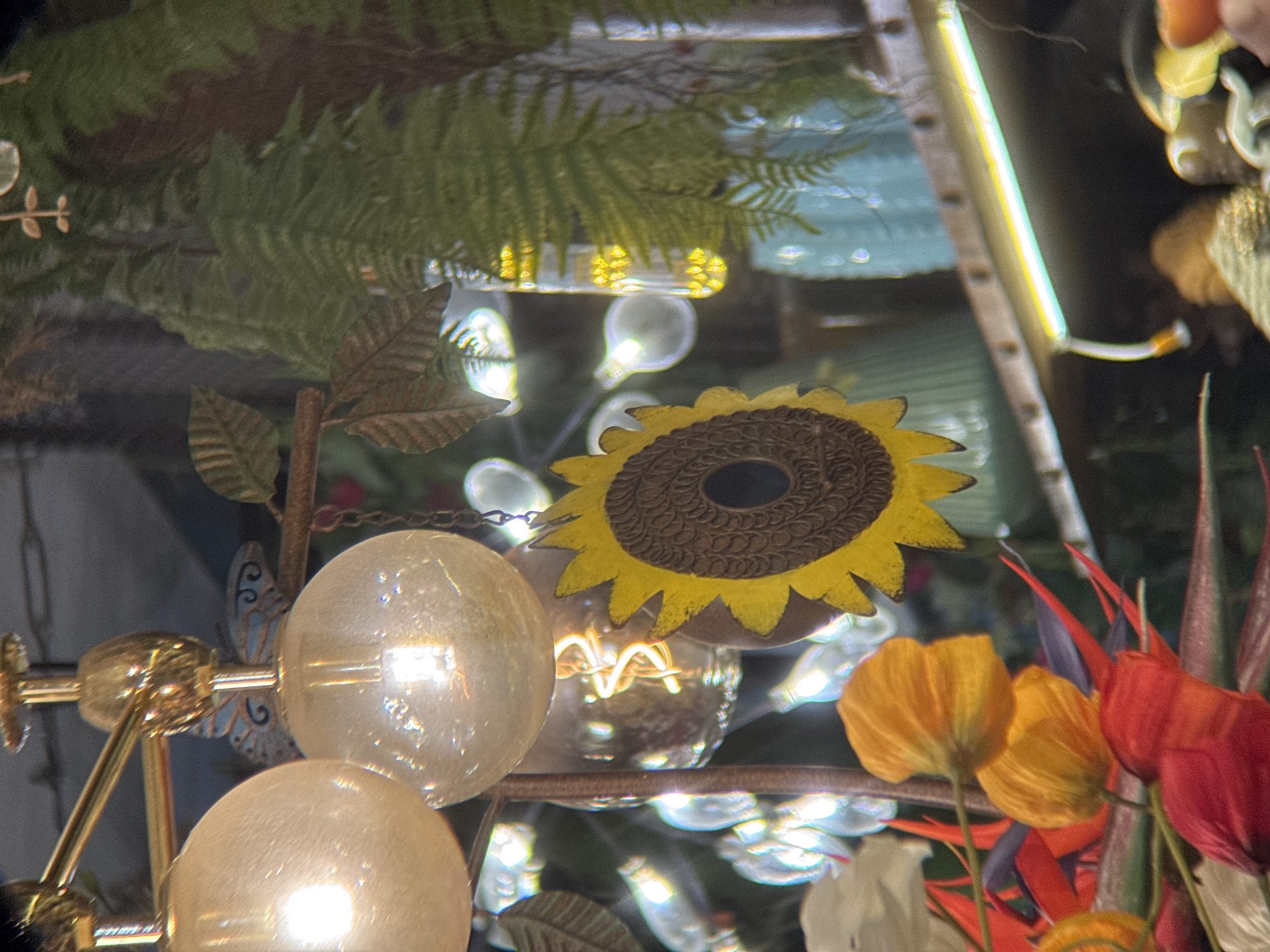 This image depicts an indoor scene, possibly a restaurant or shop, with an array of decorative elements. A prominent feature is a chandelier with large, frosted glass bulbs emitting a warm light. Dangling from the chandelier is a decorative sunflower made of metal and wood. Various artificial plants, including ferns and other greenery, are visible, adding to the decor. In the foreground, there are colorful artificial flowers, primarily in shades of orange and red. The background shows more light fixtures and some blurred objects, creating a festive and slightly cluttered atmosphere. The overall impression is one of a whimsical and elaborately decorated space. The image does not provide enough visual information to determine its specific location within Nha Trang, Vietnam.