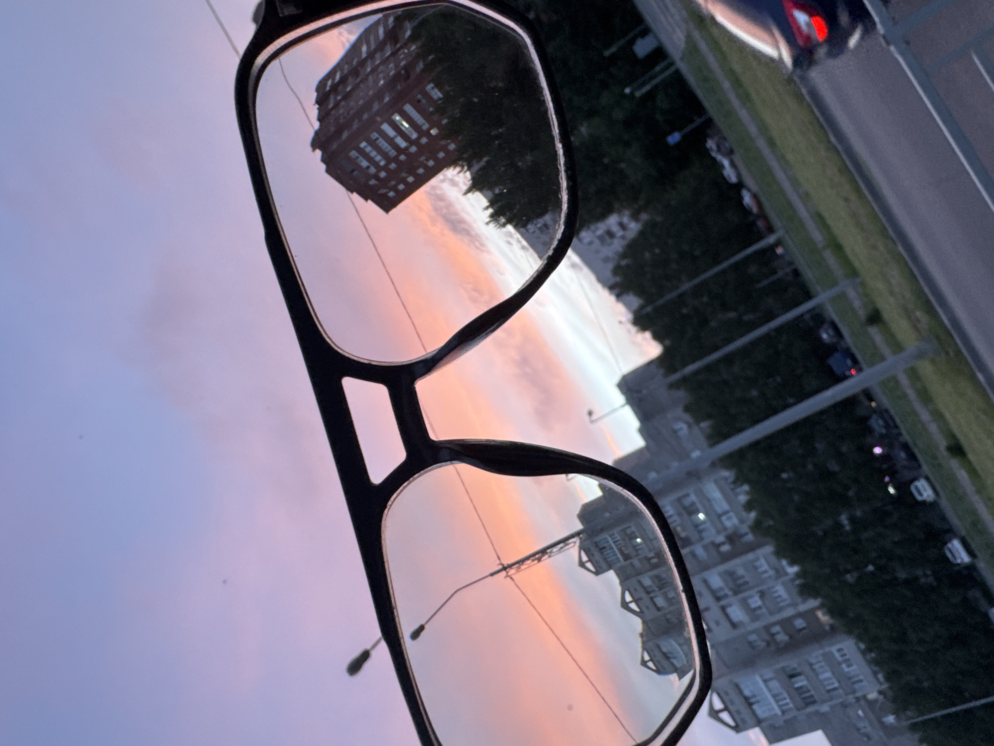 A pair of dark-framed eyeglasses is held by an unseen individual, angled to encompass an urban landscape at sunset or dusk. The sky, visible both within the lenses and around the frame, displays a pronounced color gradient, transitioning from light blue and lavender in the upper portions to vivid orange and pink hues along the horizon, indicative of a sunset. Scattered light clouds are present within the colorful sky.
Through the left lens, a multi-story, light-colored concrete residential building with visible balconies and windows is observed against the bright sky. A street lamp post, featuring a vertical pole and two horizontal light fixtures, is sharply reflected within this lens, appearing somewhat centrally located.
Through the right lens, a taller, dark red-brick multi-story building, likely residential, is depicted. Its windows reflect the bright, colorful sky. This building is presented at an oblique angle.
Beyond the eyeglasses, in the background, multiple grey, multi-story residential buildings are faintly visible, characteristic of urban architecture. A line of dark green trees separates the buildings from a paved road that crosses the lower part of the image, ascending slightly from left to right.
On the right side of the road, the rear section of a dark-colored vehicle, possibly a hatchback or compact SUV, is partially visible, with its red taillight illuminated. Further along the road and in adjacent areas, several other parked or stationary vehicles, primarily white and dark, are discernible. Numerous street lamp posts and utility poles with overhead wires are distributed across the urban background. A white metal barrier or railing extends across the bottom right foreground.
The overall scene is bathed in the soft, ambient light of either sunset or dawn, creating a stark contrast between the luminous sky and the more subdued, shadowed ground elements. The specific location is in the city of Unknown, Unknown.