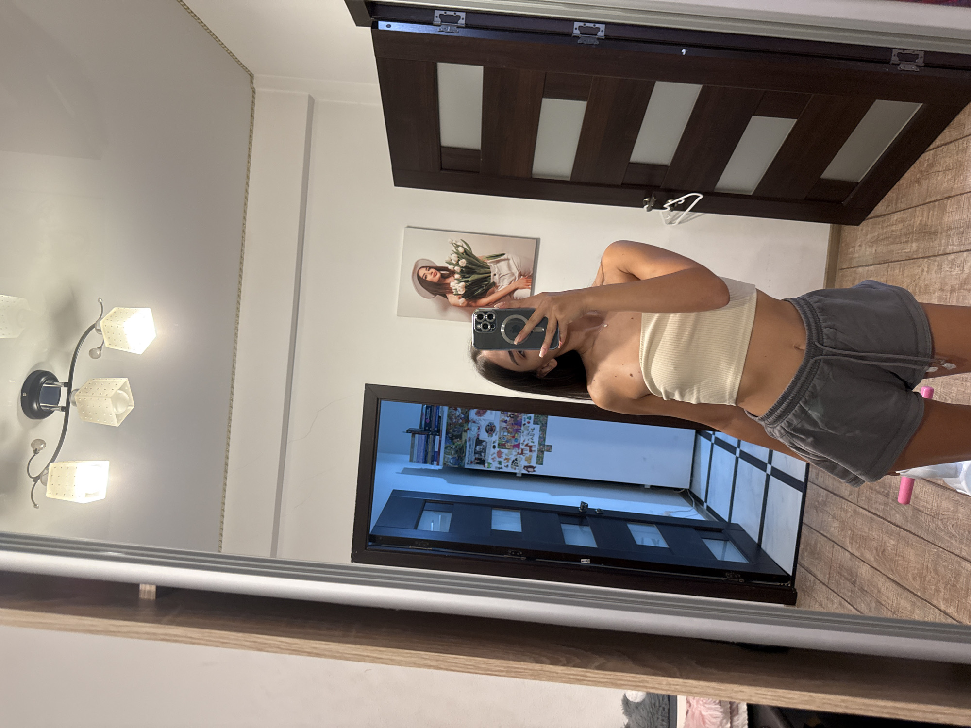 A young woman, appearing to be in her late teens or early twenties, is taking a mirror selfie in what appears to be a residential interior. She is wearing a light-colored, strapless crop top and grey shorts. Her hair is dark and falls past her shoulders. She is holding a smartphone in her right hand to capture the reflection.
The environment includes a large mirror which dominates the left side of the frame, reflecting the woman and the room behind her. To the right of the mirror, there are two dark wooden doors. One door is partially open, revealing a hallway with checkered flooring and a doorway at the end. The other door is closed and has horizontal glass panels. On the wall between the doors, there is a framed picture of a woman holding a bouquet of flowers. Above this picture, a ceiling light fixture with three square lamps is visible. The floor is made of light-colored wooden planks.
The specific location within Chișinău, Moldova, cannot be determined from the visual information provided in the image.