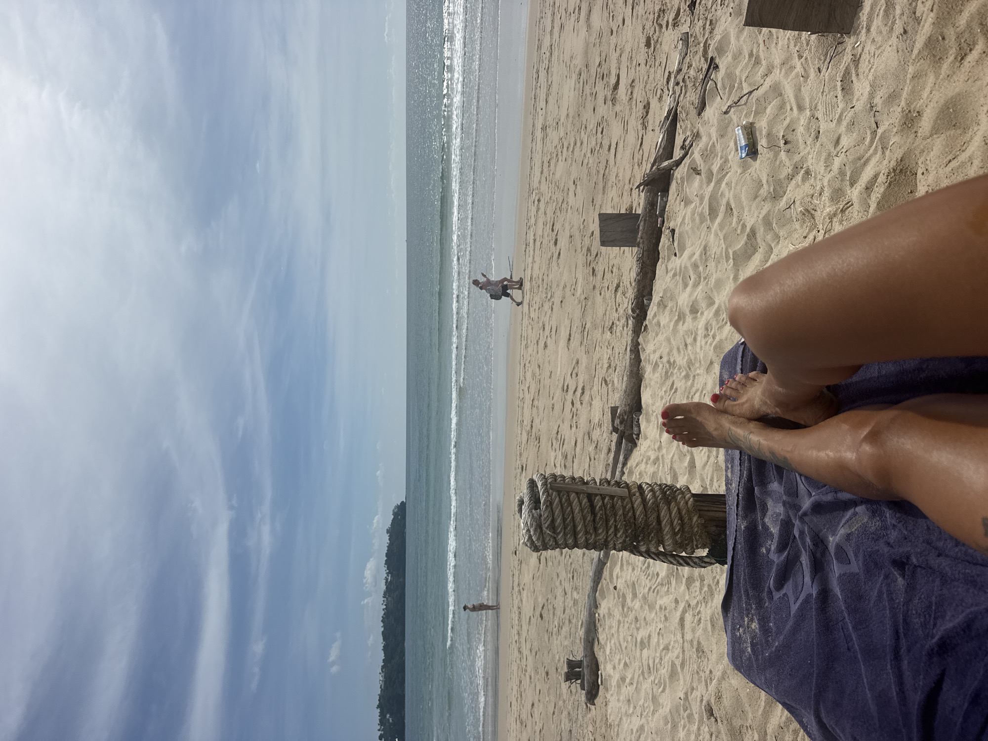 The image depicts a sun-drenched beach scene in Choeng Thale, Thailand. In the foreground, a person's legs, adorned with red nail polish, rest on a dark blue towel. The sand is a light golden color and shows the texture of ripples. A thick rope is coiled around a wooden post, situated on the sand. In the midground, the ocean meets the shore with gentle waves. Two people are walking along the water's edge, one appearing to be a child and the other an adult. Further in the background, a verdant, tree-covered hill or landmass is visible against a bright blue sky with scattered clouds. The overall atmosphere is one of a peaceful day at the beach.
