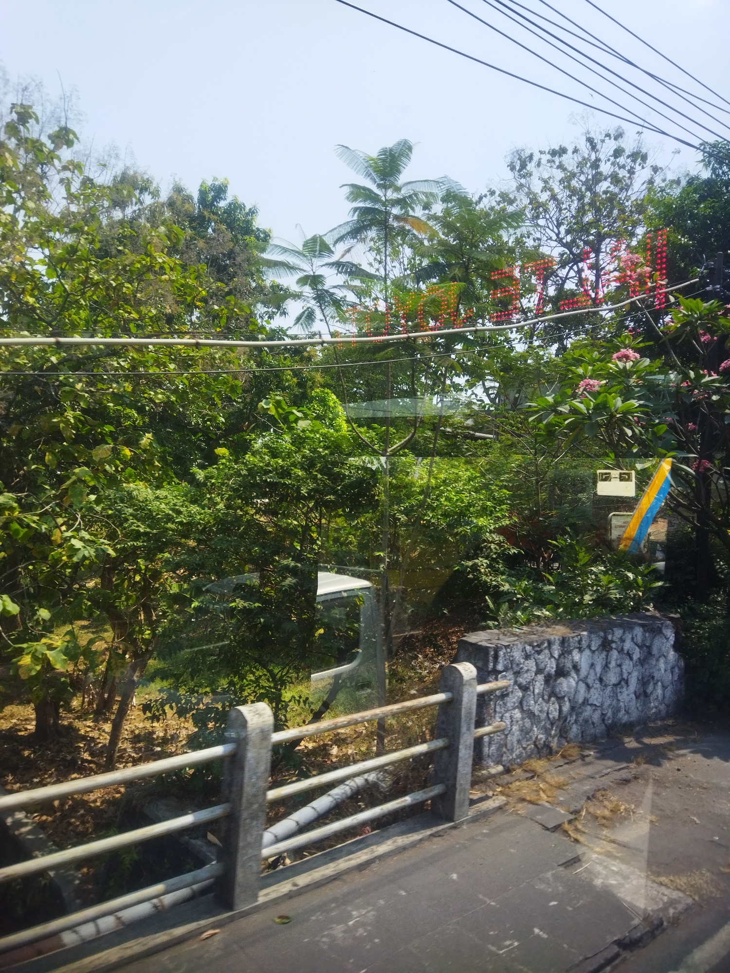 The image shows a view from a vehicle, looking out through a window at a lush green environment. In the foreground, there is a metal railing with concrete posts, a paved road surface, and a low stone wall. Behind the railing and wall, dense foliage and trees are visible. A reflection on the window shows part of a vehicle's interior. Above the trees, a series of red LED lights forming text is visible against a pale sky, along with electrical wires. The text appears to be "HOTEL". The scene suggests an outdoor setting, possibly on the outskirts of or within a town, with a focus on natural elements interspersed with man-made structures and signage. The presence of the LED sign and the road infrastructure implies an urban or semi-urban environment in Surakarta, Indonesia.