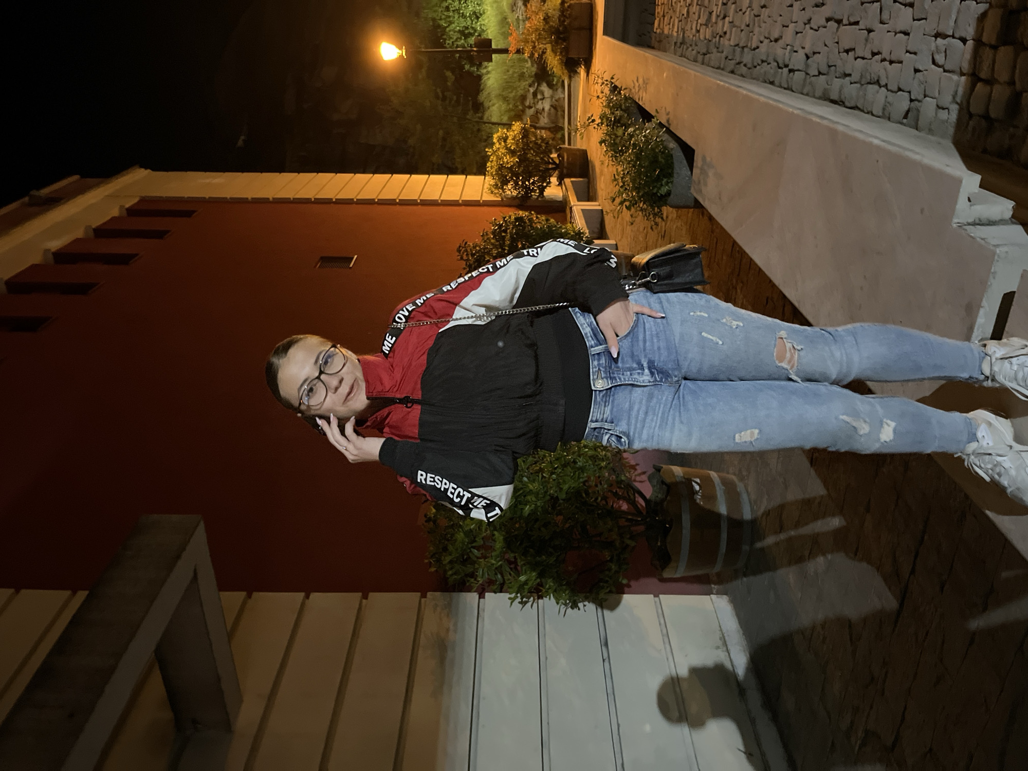 The image is taken at night in Plovdiv, Bulgaria. A young woman with glasses stands on a sidewalk adjacent to a building with a red facade. The woman is wearing a red, white, and black jacket, light blue jeans with rips, and white sneakers. She holds a phone to her ear, suggesting she is on a call, and has her left hand in her pocket. A black purse hangs from her shoulder. Behind her, plants in pots sit along the building's edge. A streetlight illuminates the scene. The sidewalk is made of cobblestones. The image is tilted, with the top of the building appearing at the bottom right.