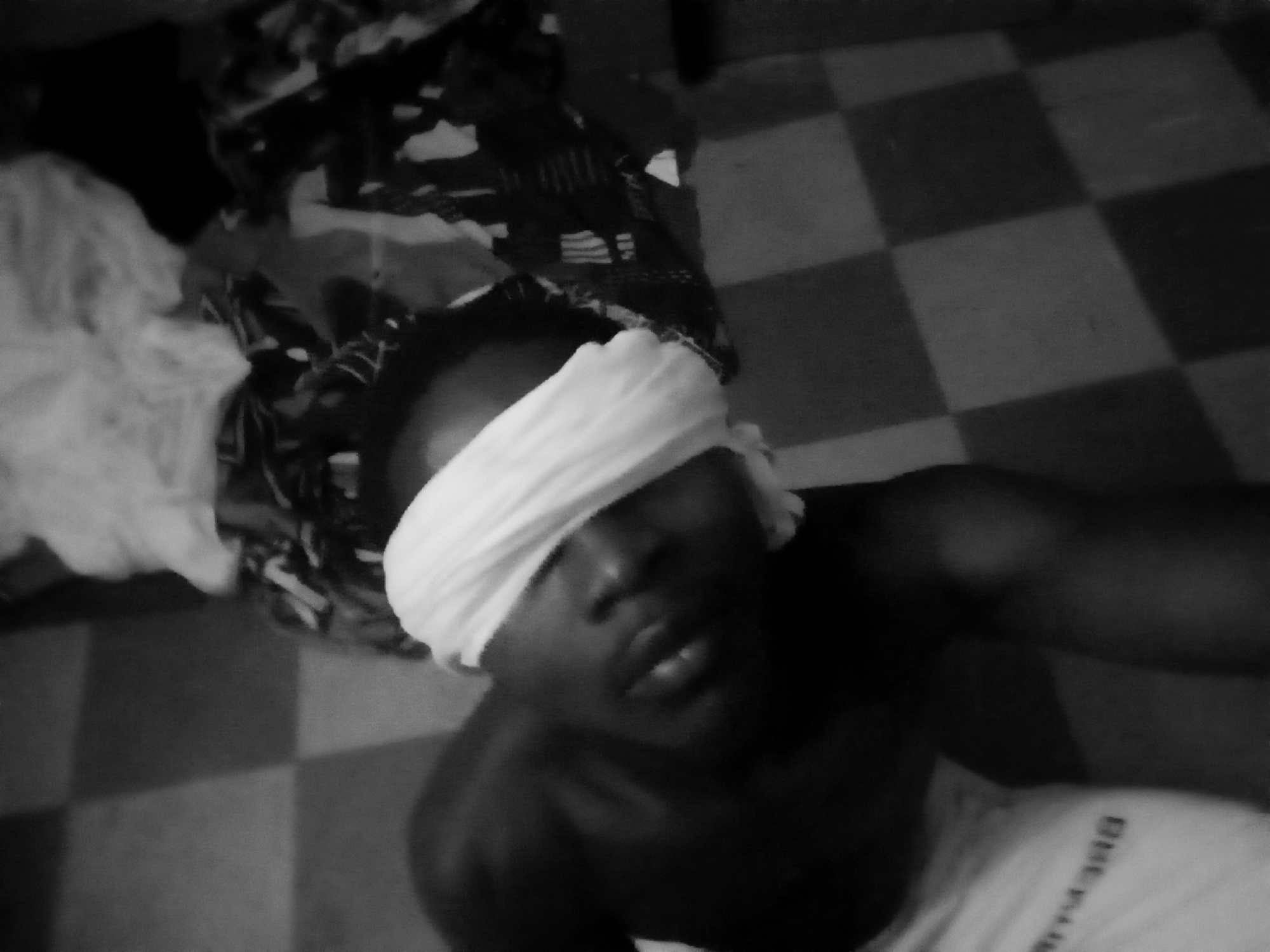This black and white photograph features a person, appearing to be male and with dark skin, positioned on a tiled floor. A white cloth is wrapped tightly around their head, completely covering their eyes as a blindfold. The person's head is tilted slightly upwards, and their mouth is slightly open, giving a neutral or vulnerable expression. They are shirtless, with a portion of a white garment visible at the bottom of the frame. On this white garment, faint and barely legible text, possibly "BHE-MAN" or similar, can be seen in the bottom right corner.
The setting appears to be indoors, possibly a room in Nkpor, Nigeria, as provided by the context. The floor is covered with a black and white checkerboard tile pattern. In the upper left background, a pile of assorted dark and light patterned fabrics, possibly clothes or blankets, lies somewhat disheveled. The overall lighting is dim, contributing to a somber or intimate mood. The image has a grainy texture and is slightly blurry. The activity depicted appears to be a staged or posed moment, emphasizing vulnerability or quiet introspection through the act of being blindfolded.