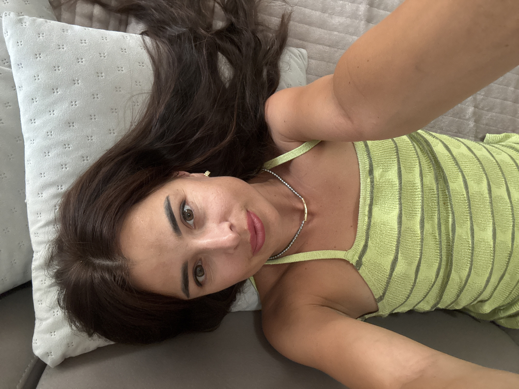 A woman with long, dark hair is lying on a gray couch with a light green, striped top and a pillow. Her head is tilted back, and she is looking up at the camera. Her right arm is raised, and her hand is out of frame. She is wearing a necklace and earrings. The scene appears to be indoors, possibly in a bedroom or living room.