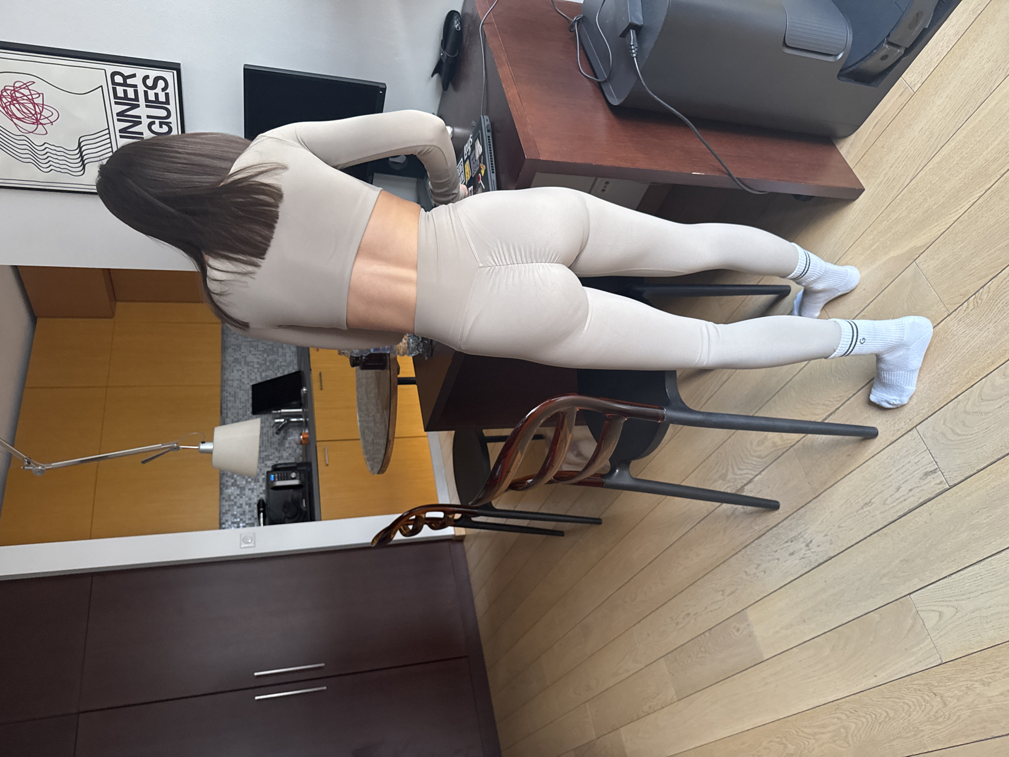A woman is captured mid-exercise, performing a plank in an indoor setting, viewed from behind. She has long, straight dark hair and is dressed in a light beige or pale taupe long-sleeved crop top and matching high-waisted leggings. Her feet wear white athletic socks adorned with black stripes and a small green emblem. Her body is held in a straight line, parallel to the light-colored wooden floorboards.
The scene depicts a modern room, likely an apartment in Warsaw, Poland. In the immediate vicinity of the woman, a dark brown wooden desk or table is visible, holding a black computer monitor, a laptop, and various cables. A contemporary chair with dark legs and a brown, sculptural backrest sits beneath the desk. To the left, a dark brown cabinet with two silver handles is present.
The background reveals a kitchen area equipped with light wood cabinets, a grey tiled backsplash, and an extended white adjustable desk lamp with a conical shade. On the wall above the desk, a framed poster features abstract red scribbles on a wavy white background with the partial text "INNER GUES" visible. The room is brightly lit, suggesting it is daytime. The activity is clearly a fitness exercise, demonstrating focus and physical exertion.