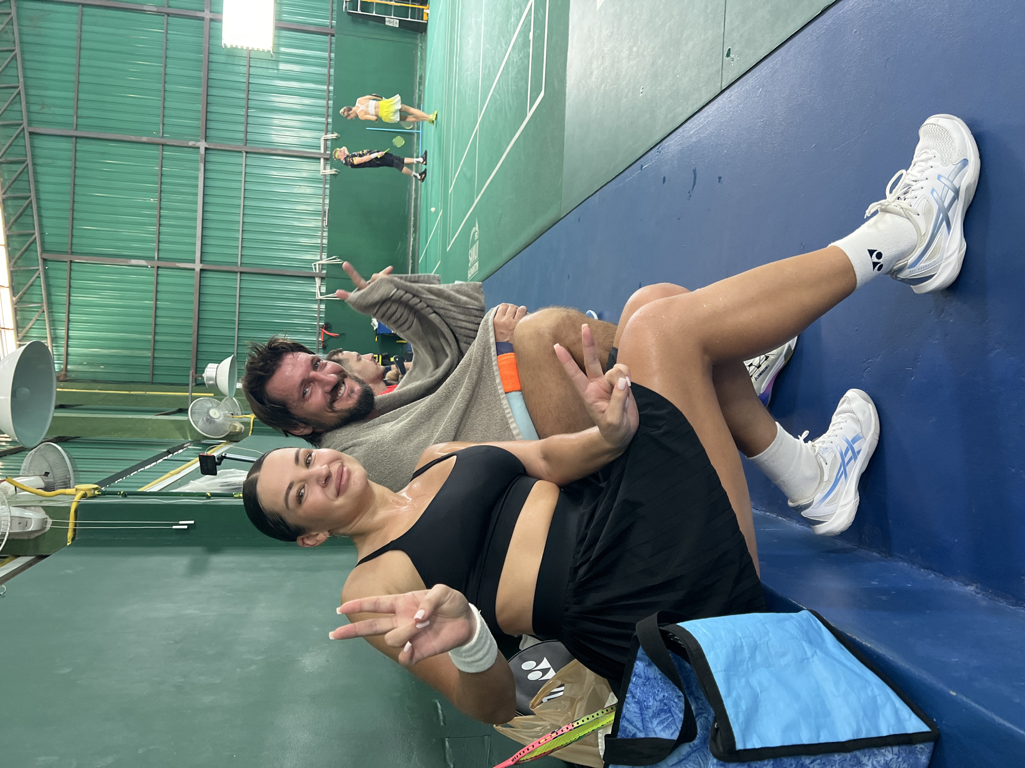 In an indoor badminton court in Ban Bang Rak, Thailand, a Caucasian woman with dark hair pulled back and a man with dark hair and a beard sit on a blue bench. Both are smiling and holding up peace signs with their hands. The woman wears a black sports bra, a black pleated skirt, white socks, and white and blue athletic shoes. The man wears a grey sweater over an orange and blue-striped shirt and blue shorts. A grey towel is draped over his shoulders and the bench. Next to the woman are a blue bag and a badminton racket. In the background, two people are playing badminton on the court. The court has green flooring with white lines. The walls of the court are dark green. Lighting fixtures and ceiling fans are visible overhead.