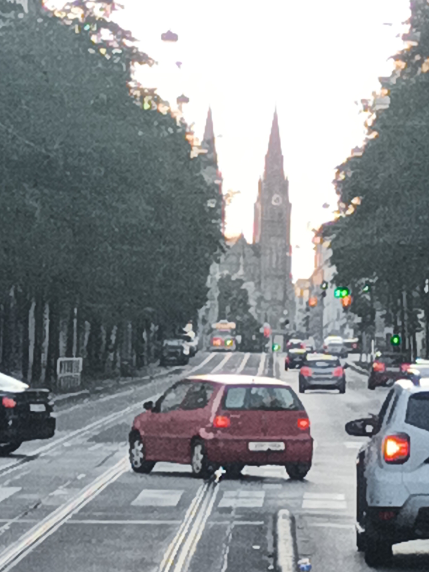 This is a photograph taken in Prague, Czechia, featuring a street scene with a prominent church or cathedral in the background. The architecture suggests a historical European city. The foreground shows a street with tram tracks, indicating that trams are a mode of public transport in this location. Several cars are visible on the road, suggesting moderate traffic. The lighting suggests it might be late afternoon or early evening, with the sun low in the sky. The overall impression is of an urban environment with a blend of historical and modern elements. The image is somewhat blurry, which could be due to the camera's focus or motion.