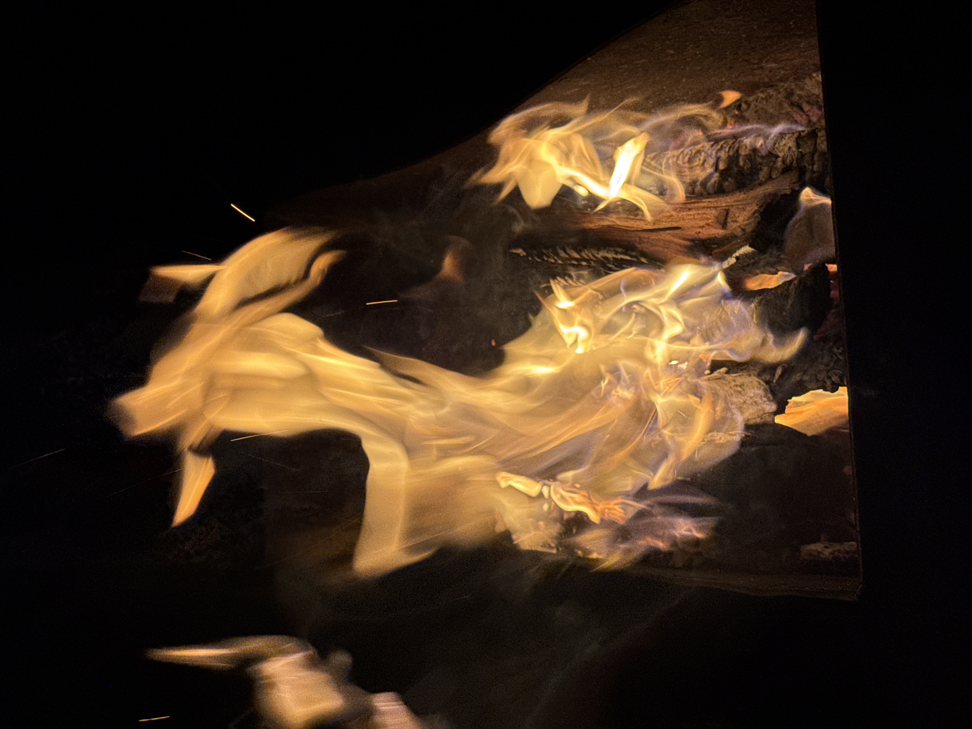 The image shows flames in a fire pit at night. The fire pit is made of dark metal. The flames are predominantly yellow and orange, with some hints of blue. Charred wood is visible within the flames. The background is dark and out of focus. The location in the city of Mayak, Russia is not evident in the image.