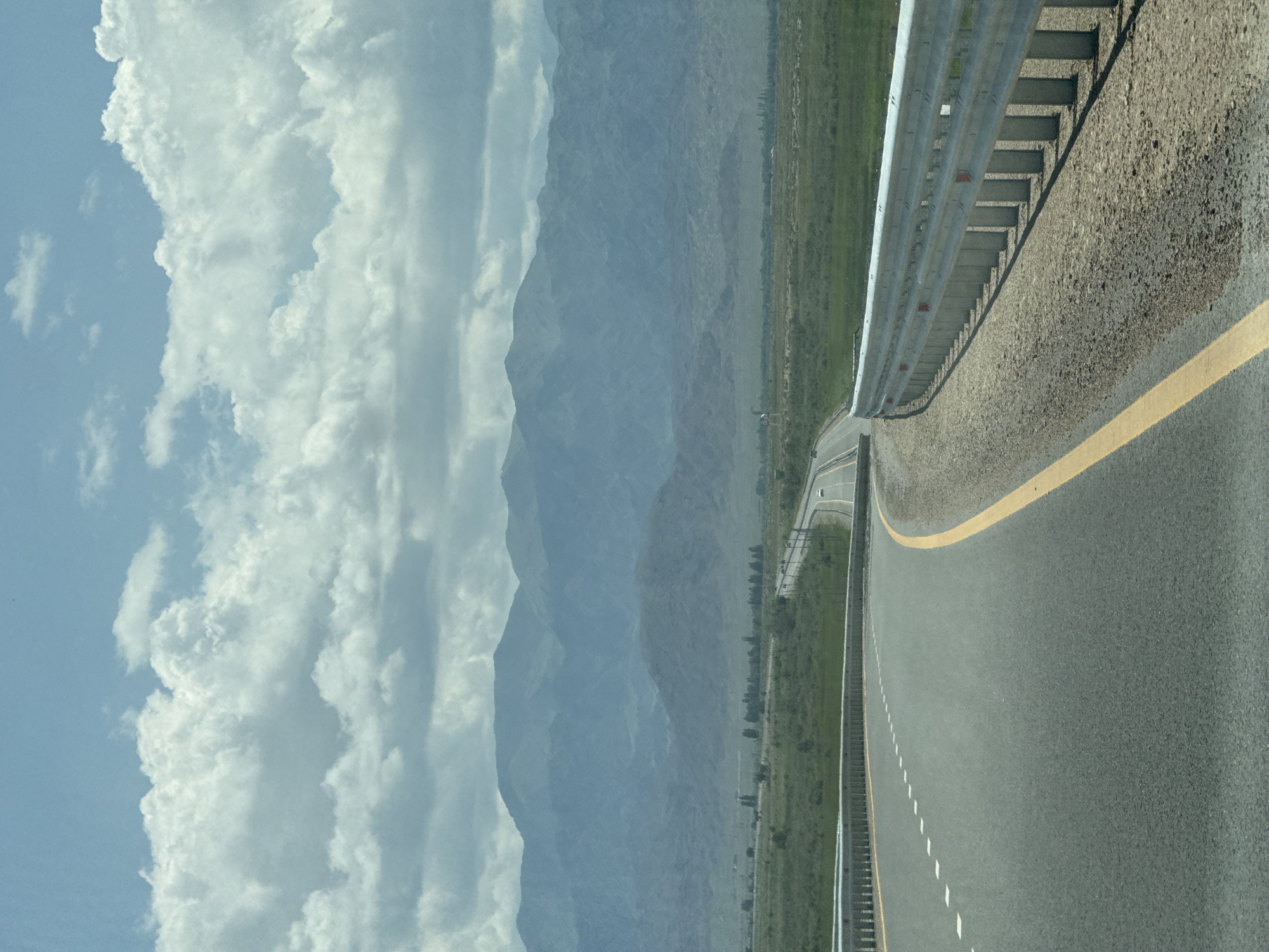 The image depicts a scenic view of a highway curving through a vast, grassy plain towards a range of distant, hazy mountains. The sky is filled with dramatic, white cumulus clouds against a pale blue backdrop. The highway itself is paved and has a yellow dividing line. A silver metal guardrail runs along the right side of the road. In the distance, a few cars can be seen on the highway, and a sparse line of trees borders the plain. The overall impression is one of open space and natural beauty, with the highway acting as a pathway through the landscape. While the specific location within Balykchy, Kyrgyzstan, is not explicitly identifiable from the visual elements alone, the scene is consistent with the geographical characteristics of the region, which features large open plains and mountainous terrain.