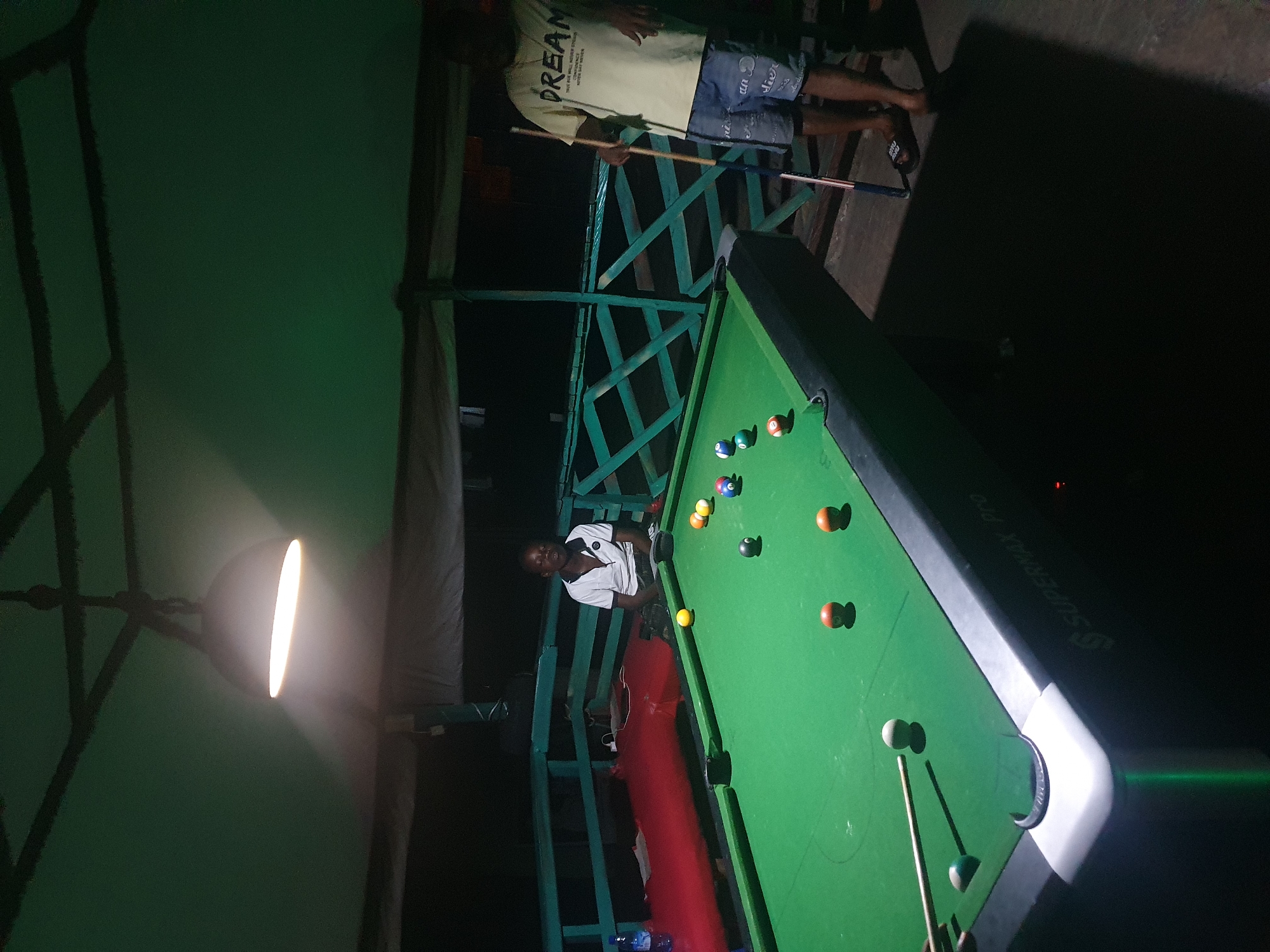 In Lokoja, Nigeria, a dimly lit outdoor space hosts a billiards game. A green-felt pool table with several balls scattered across it occupies the foreground. A young person in a white polo shirt is seated near the table, facing away from the camera. To the right, another person stands holding a cue stick, wearing a yellow t-shirt with the word "DREAM" printed on it and denim shorts. The area is covered by a green awning, with a hanging light fixture illuminating the scene. Wooden railings and a red-covered surface are visible in the background.