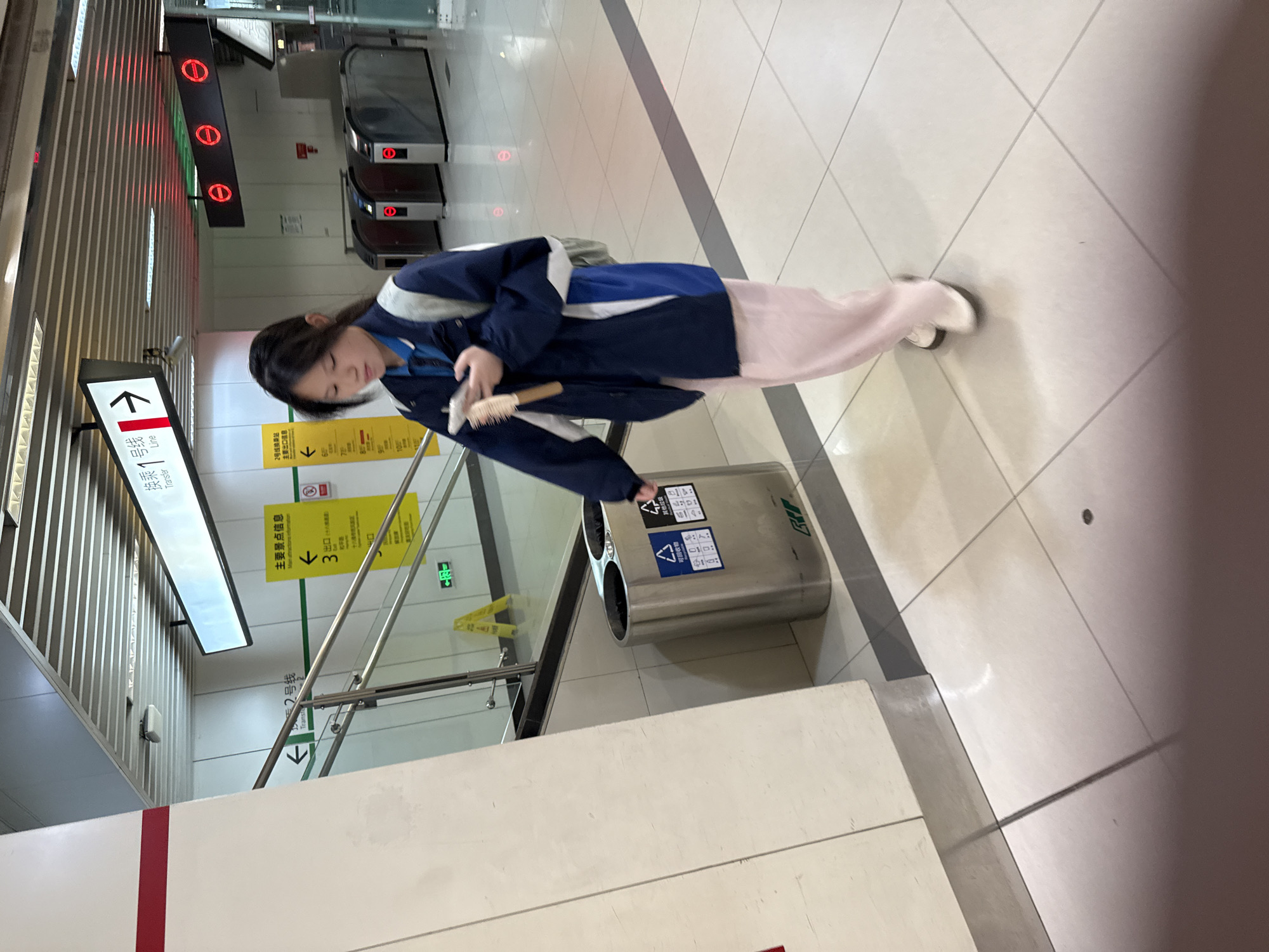 The image depicts a young woman in a public transportation setting, likely a subway station in China. She is wearing a blue jacket and light pink pants and appears to be disposing of a hairbrush in a public recycling bin. The bin is stainless steel with stickers indicating different recycling categories. The surrounding environment includes a brightly lit station interior with tiled flooring, informational signs in Chinese, and escalators in the background. The signs indicate exits and transfer information for Line 2 and Line 1, suggesting the station is a transfer point. The specific location is likely in Yuzhong District, China, based on the signage and overall appearance.