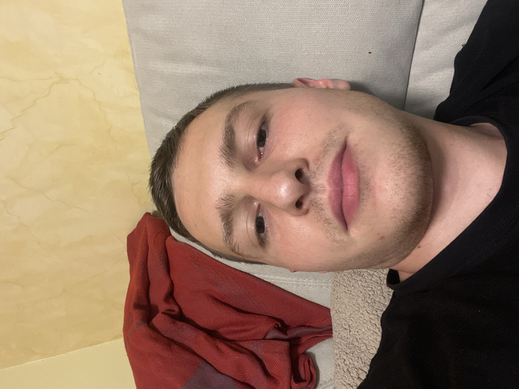 A close-up, slightly rotated portrait shot shows a Caucasian male lying on a light beige, textured surface, possibly a couch or bed. A light gray pillow rests behind his head. The man is wearing a black T-shirt. To the left, a red, folded cloth is visible. The background features a wall with a marbled yellow pattern. There is no visible information to indicate this scene is in Burgas, Bulgaria.