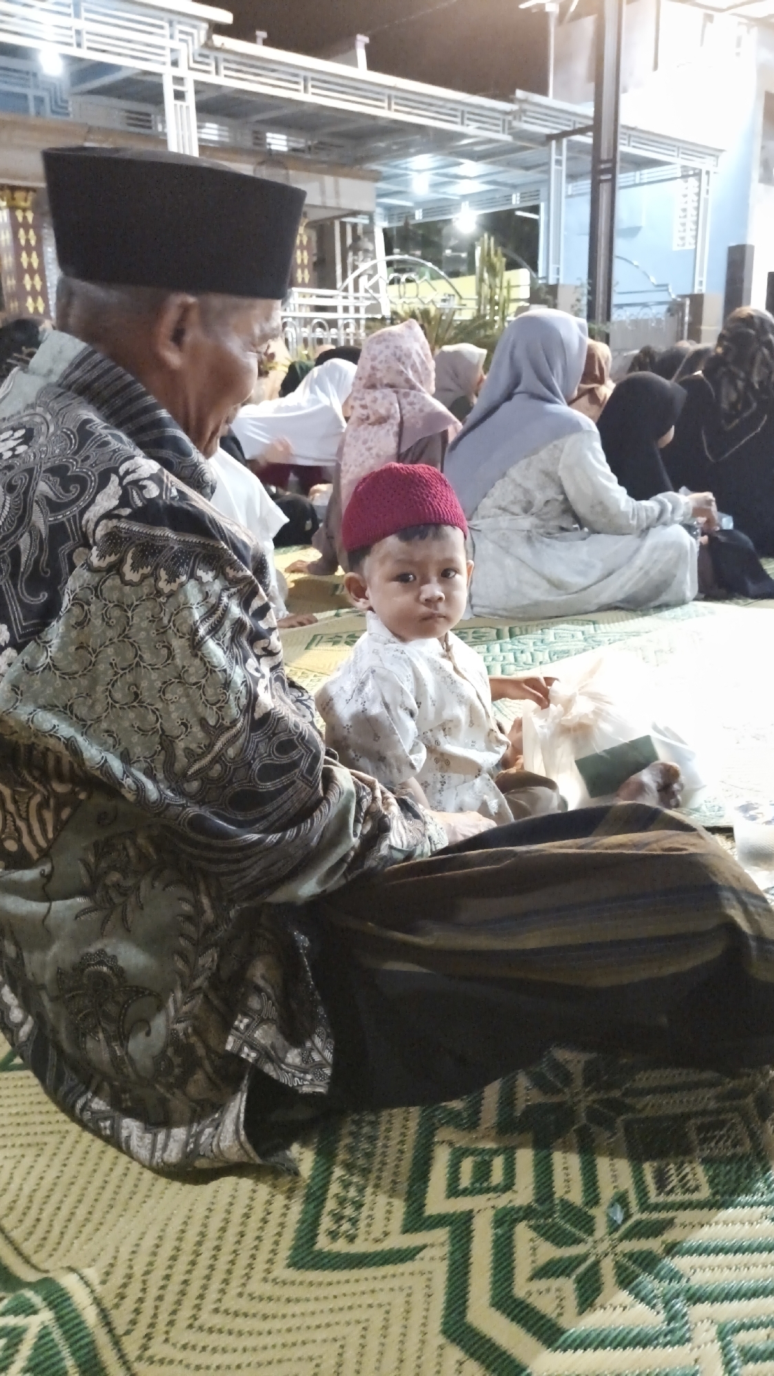A group of people, likely attending a religious or cultural gathering, are seated on a patterned mat indoors. In the foreground, an elderly man wearing a black skullcap and a traditional patterned garment is seated, with his left side facing the camera. Beside him, a young boy with a red cap sits facing the camera, wearing a light-colored garment. The boy is holding a clear plastic bag. Other individuals are visible in the background, some wearing hijabs. The environment appears to be a covered outdoor space, with a corrugated metal roof structure visible above. The scene is likely taking place in Warulor, Indonesia, based on the context.