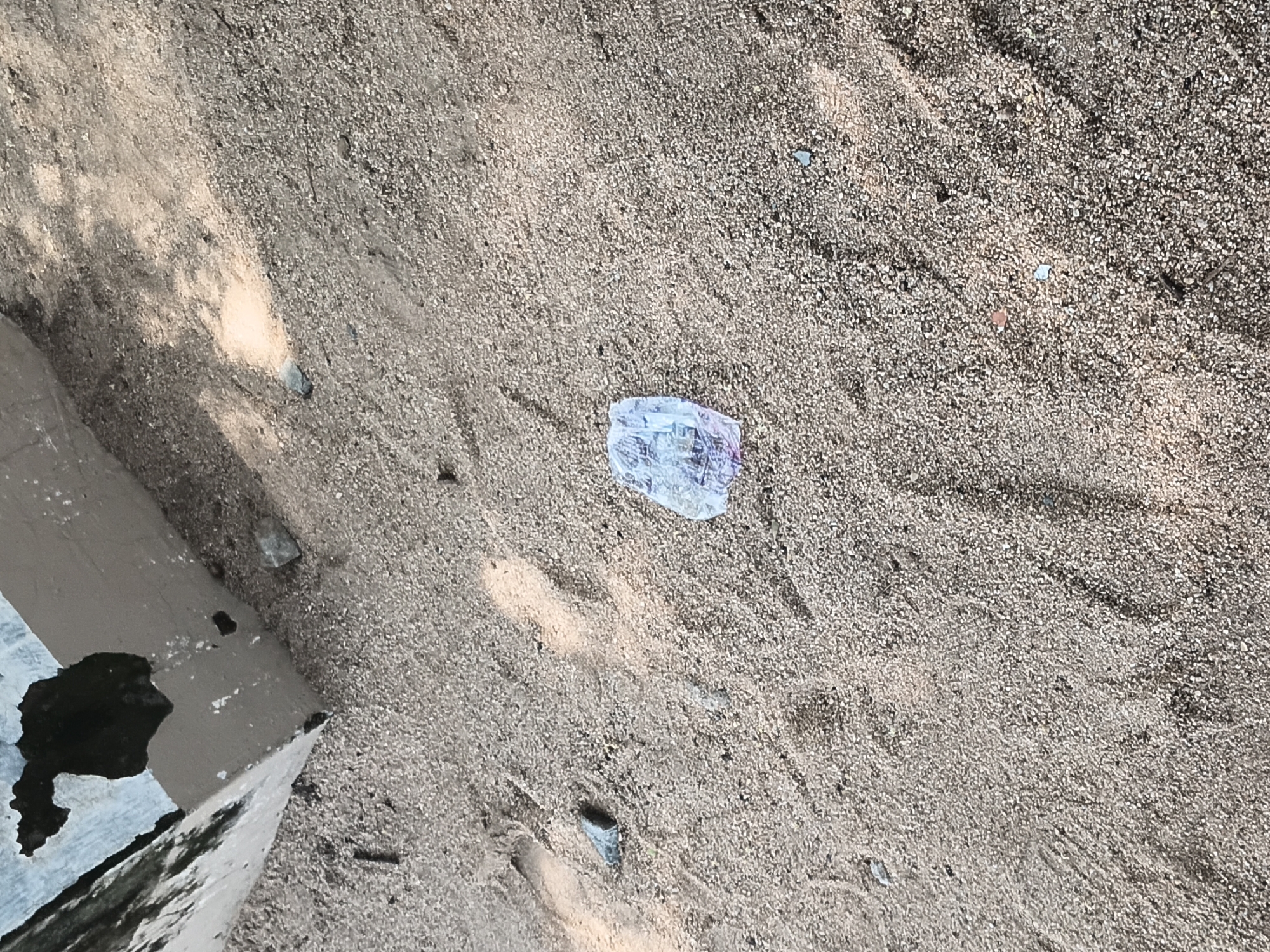 A plastic wrapper lies on a sandy ground next to a concrete structure. The wrapper is crumpled and translucent, with some purple and pink colors visible within. The concrete structure appears weathered and partially broken, with dark patches on its surface. The sandy ground is coarse-grained and shows evidence of footprints. The scene is likely located in Jalingo, Nigeria, a city known for its sandy terrain and developing infrastructure.